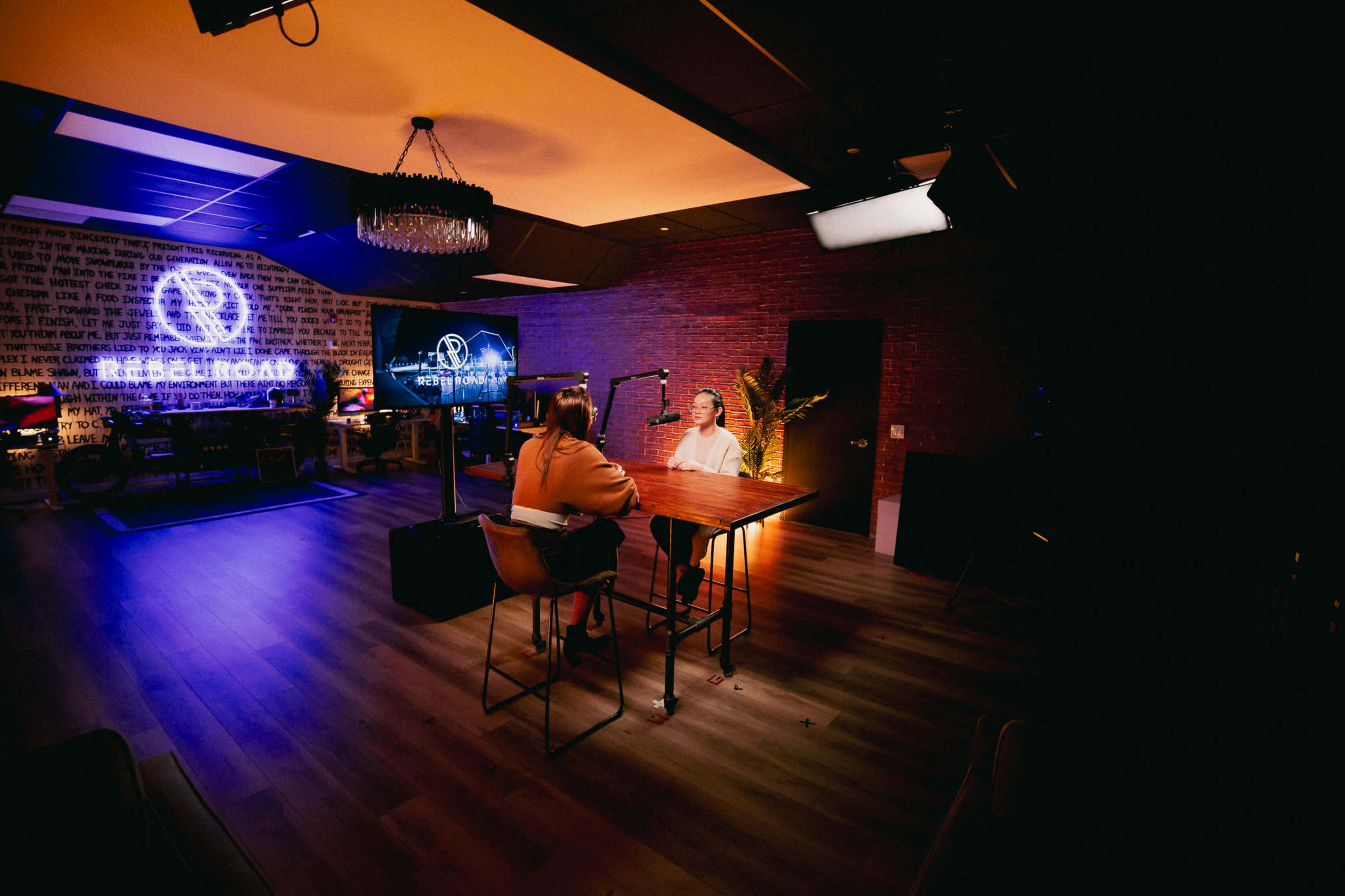 A podcast recording studio features two individuals seated at a table with microphones, surrounded by decorative lighting and screens displaying a logo.
