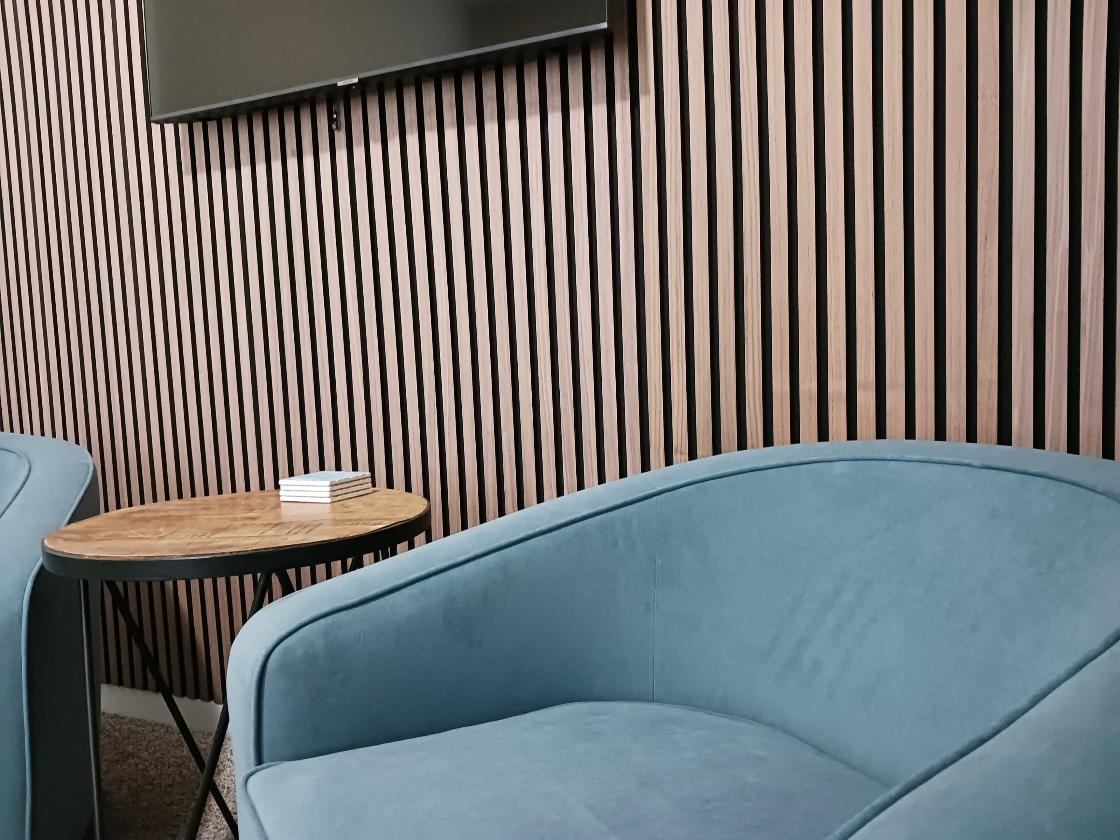 The image shows a room with a blue upholstered chair positioned beside a round wooden table, against a wall featuring vertical wooden slats.