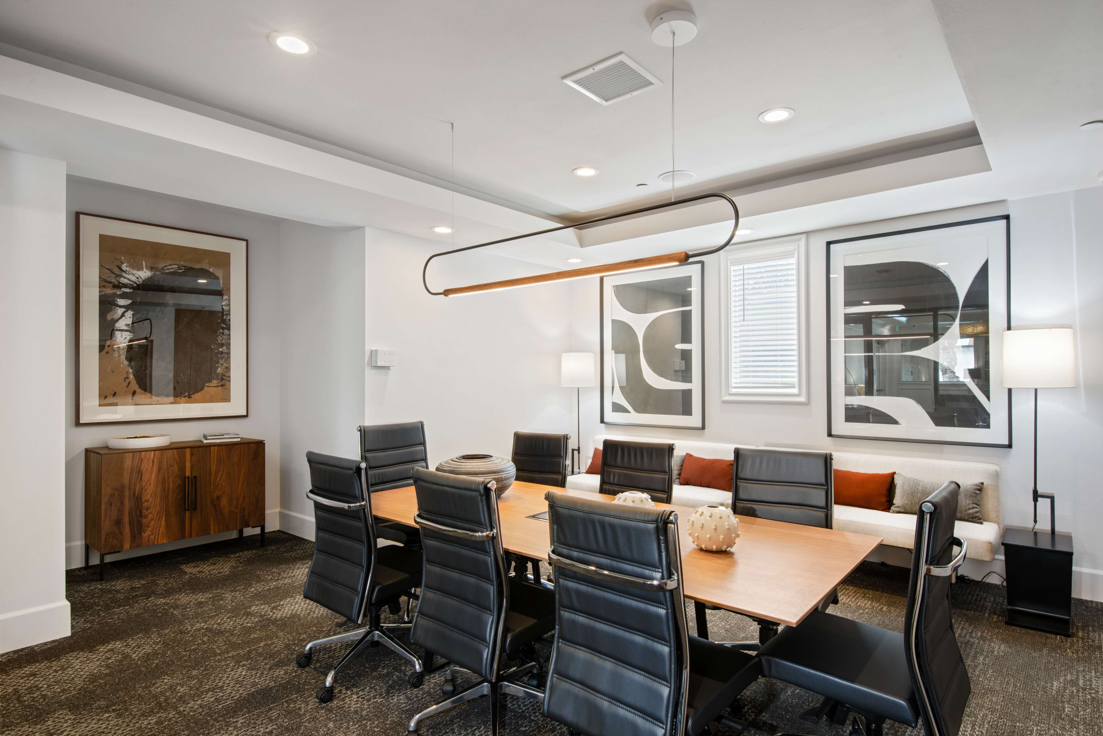 A modern conference room features a long wooden table surrounded by black chairs, with abstract art on the walls and soft seating along one side.