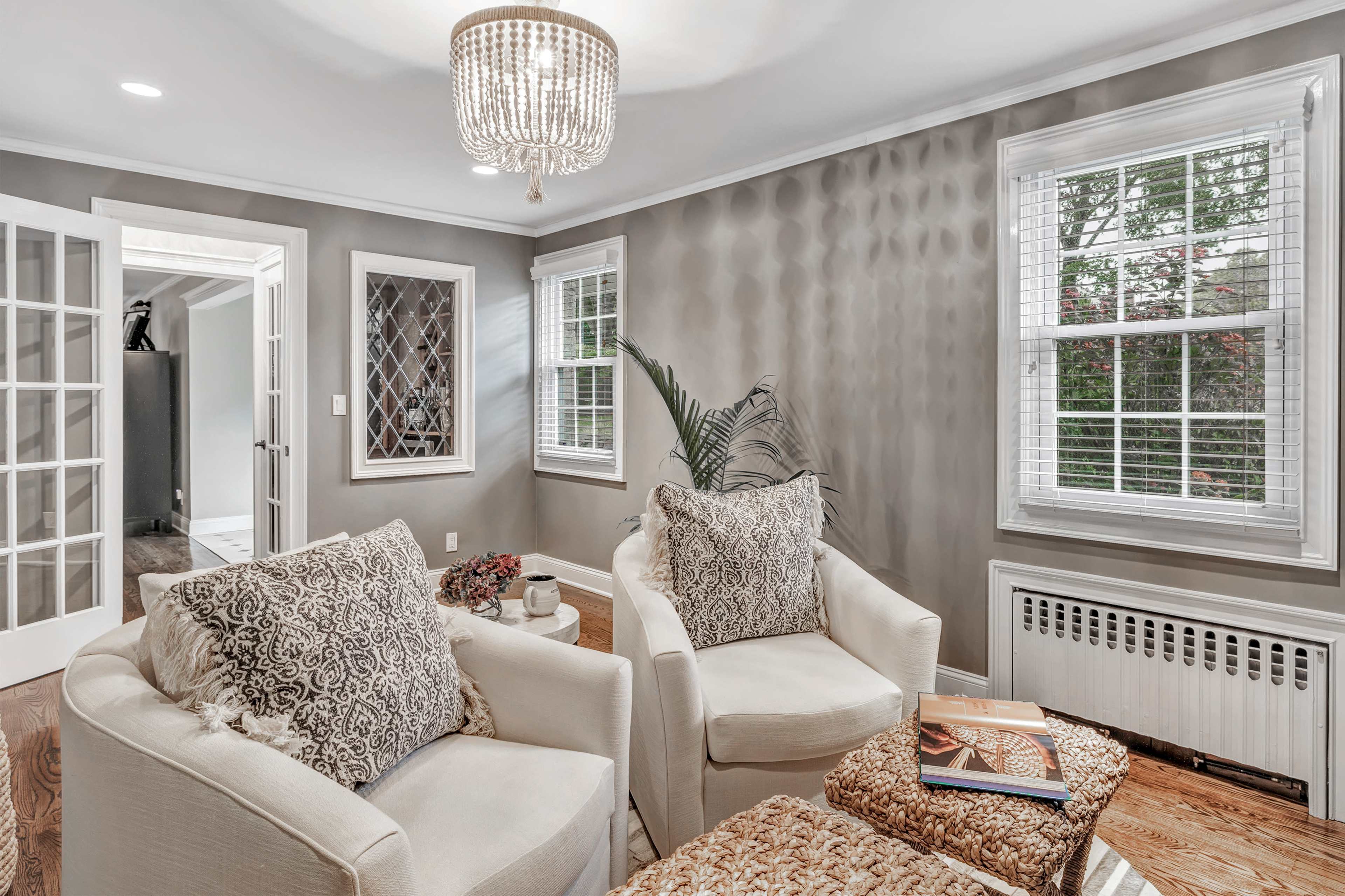 The image shows a living room with two white armchairs, a coffee table with books, and a chandelier, featuring large windows with white blinds and a textured wall.