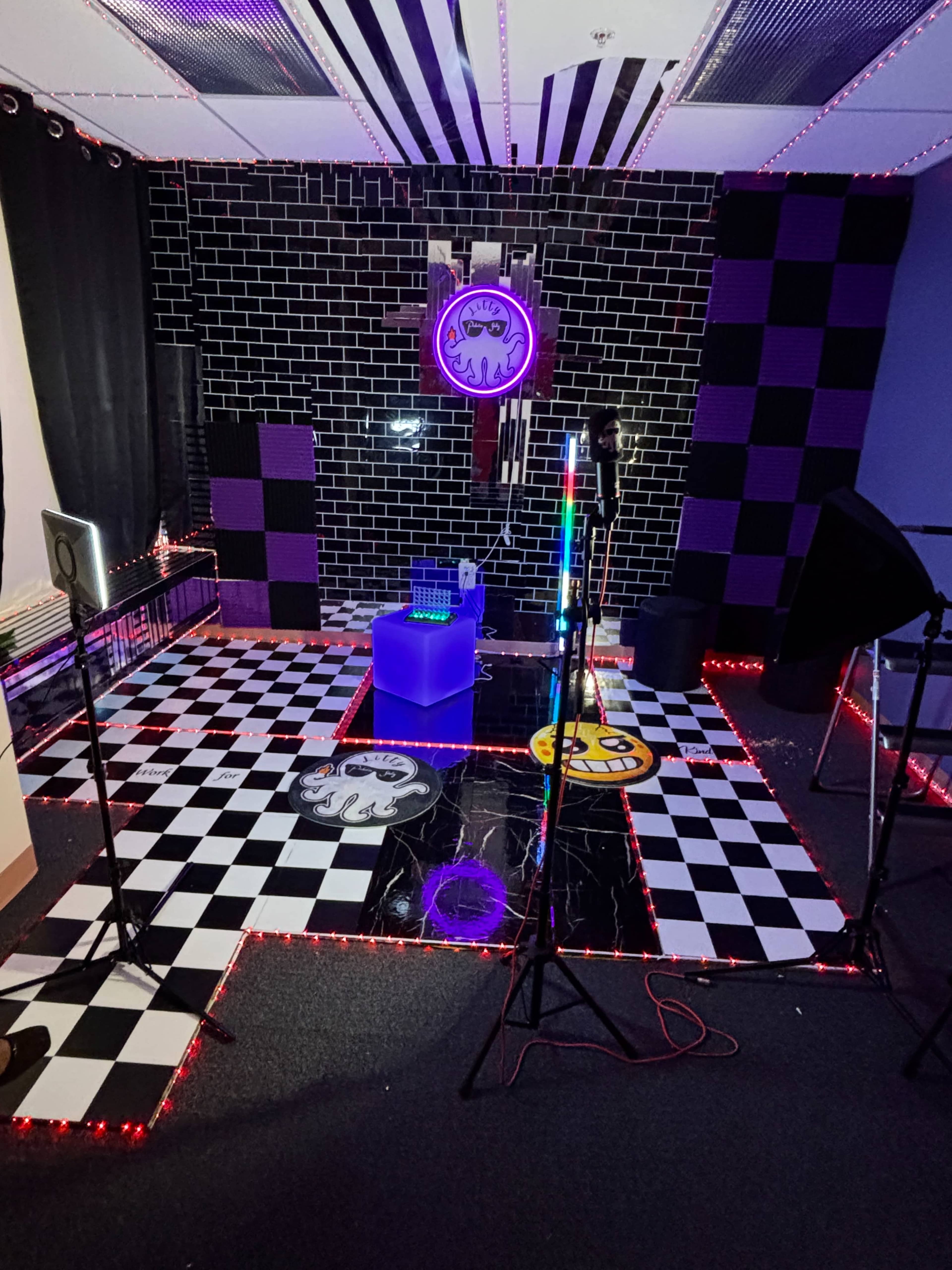 The image shows a creatively designed room with a black and white checkered floor, neon lights, and various props arranged against a dark tiled wall.