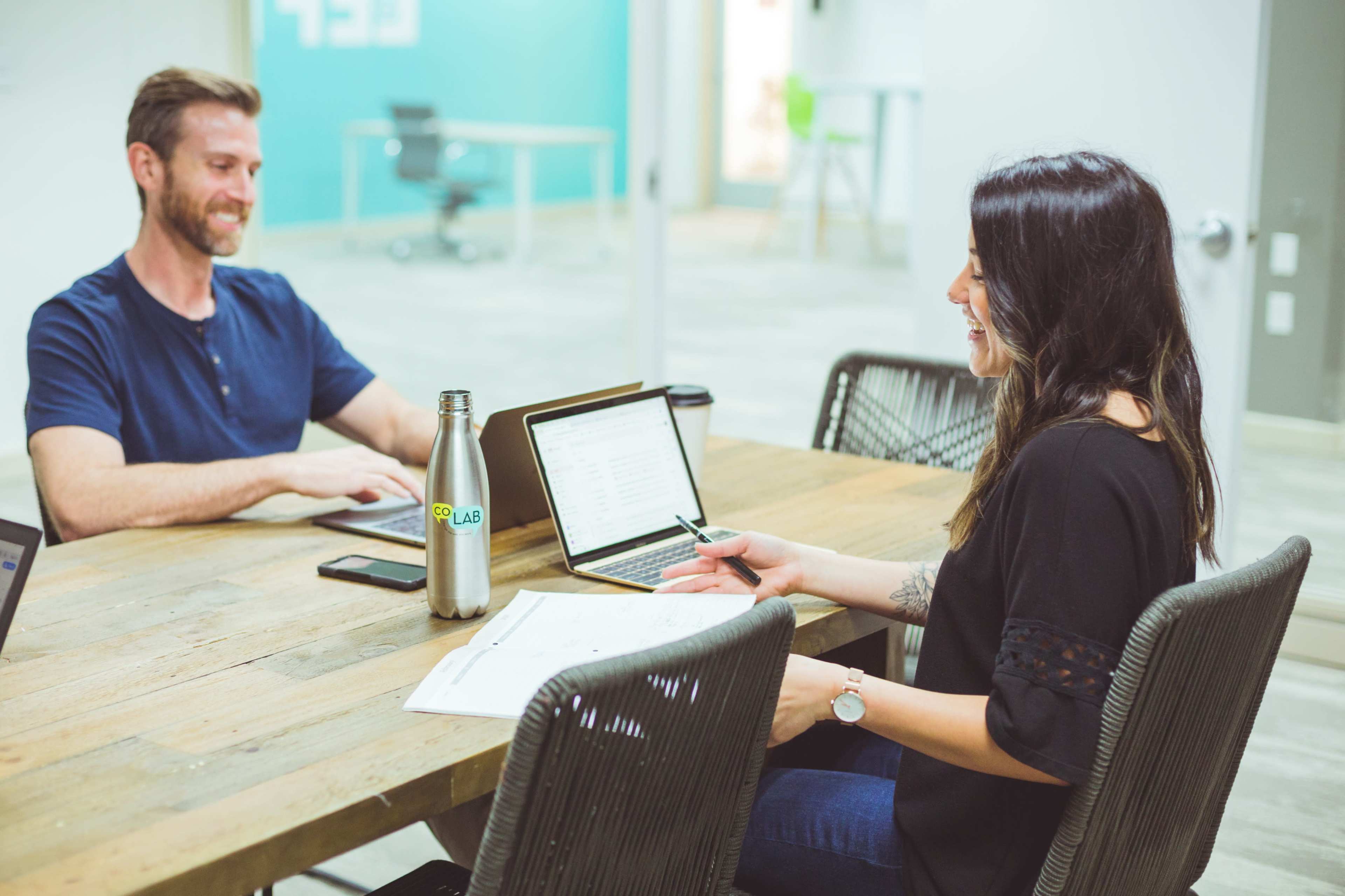 Two people sit at a wooden table in a modern office, engaged in conversation while using a laptop and taking notes.