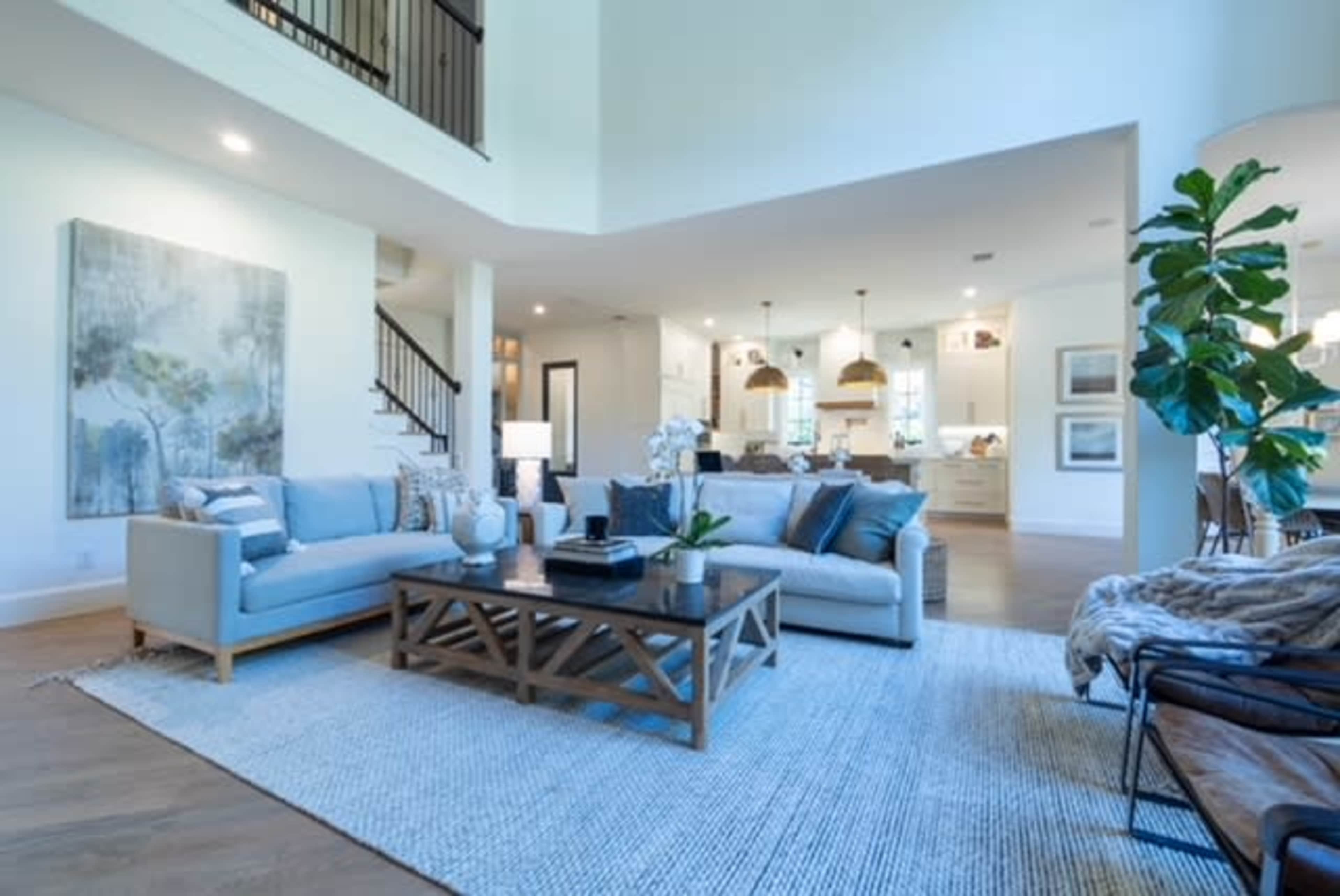 A modern living room features a light blue sofa, a wooden coffee table, and a large rug, with an open view to a kitchen area in the background.
