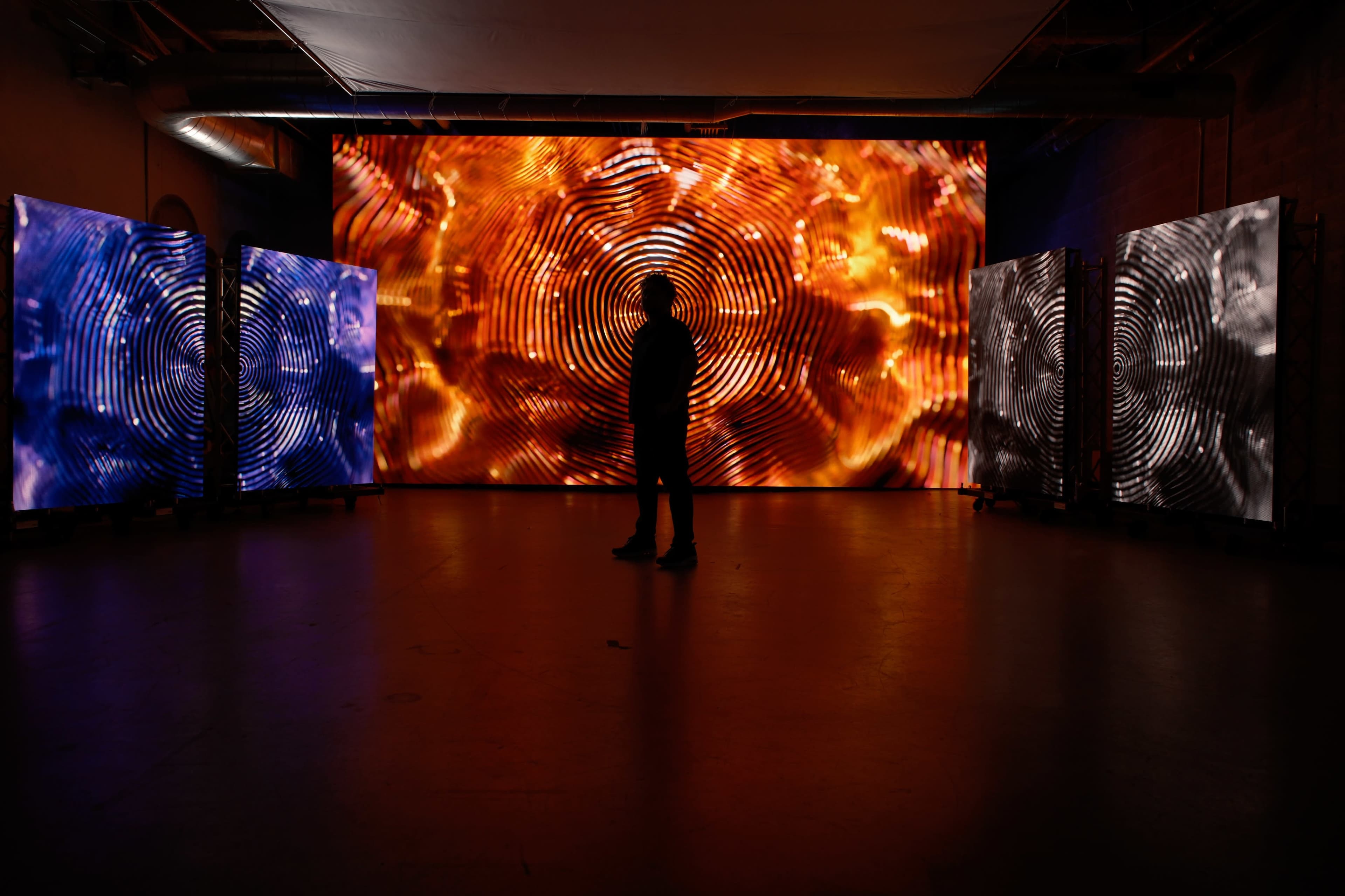 A person stands silhouetted in a dimly lit room, facing a large screen displaying vibrant, swirling patterns, flanked by two smaller screens with contrasting visuals.