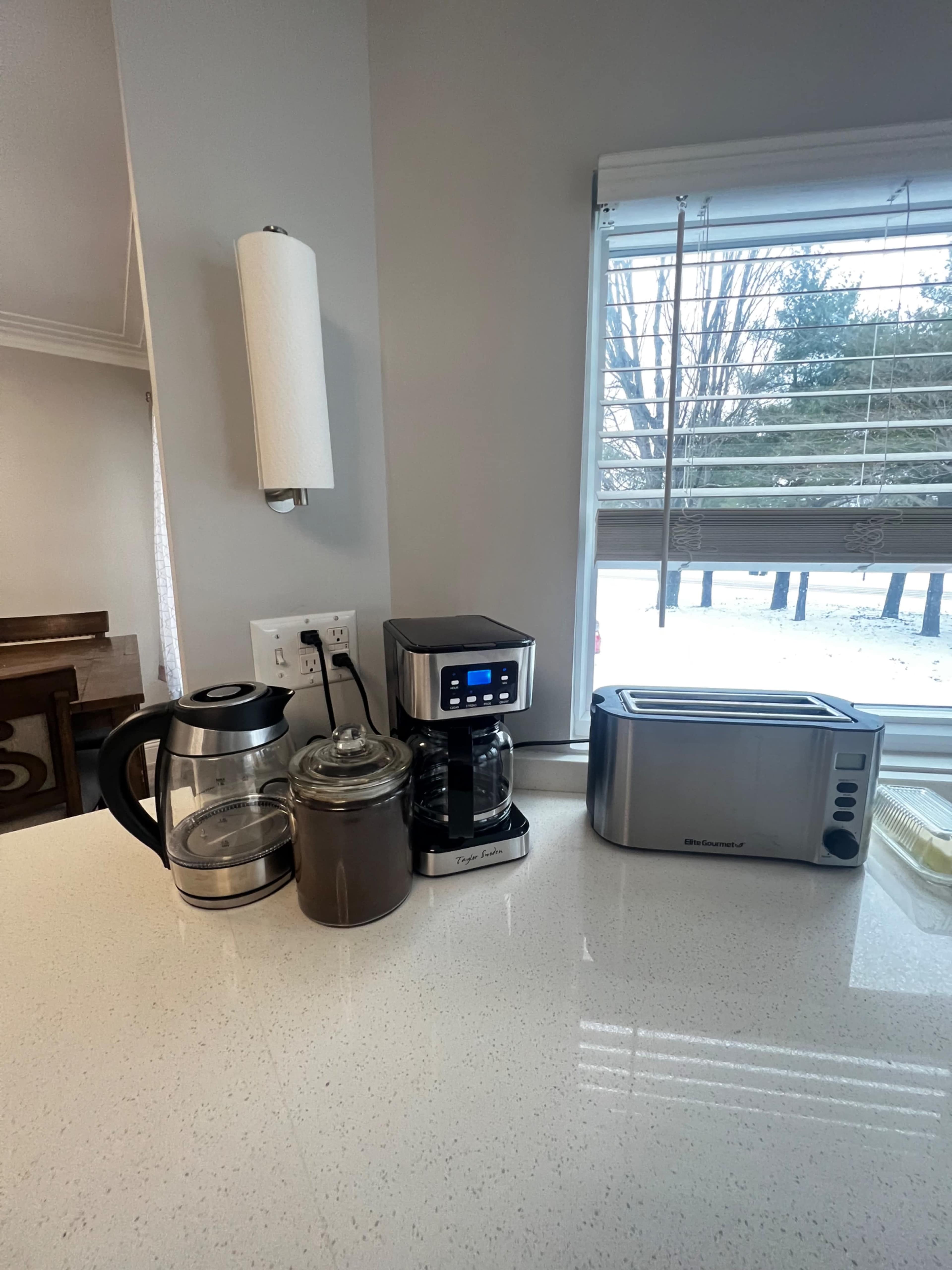 A countertop features a coffee maker, kettle, and toaster, set against a window with a view of the outdoors.