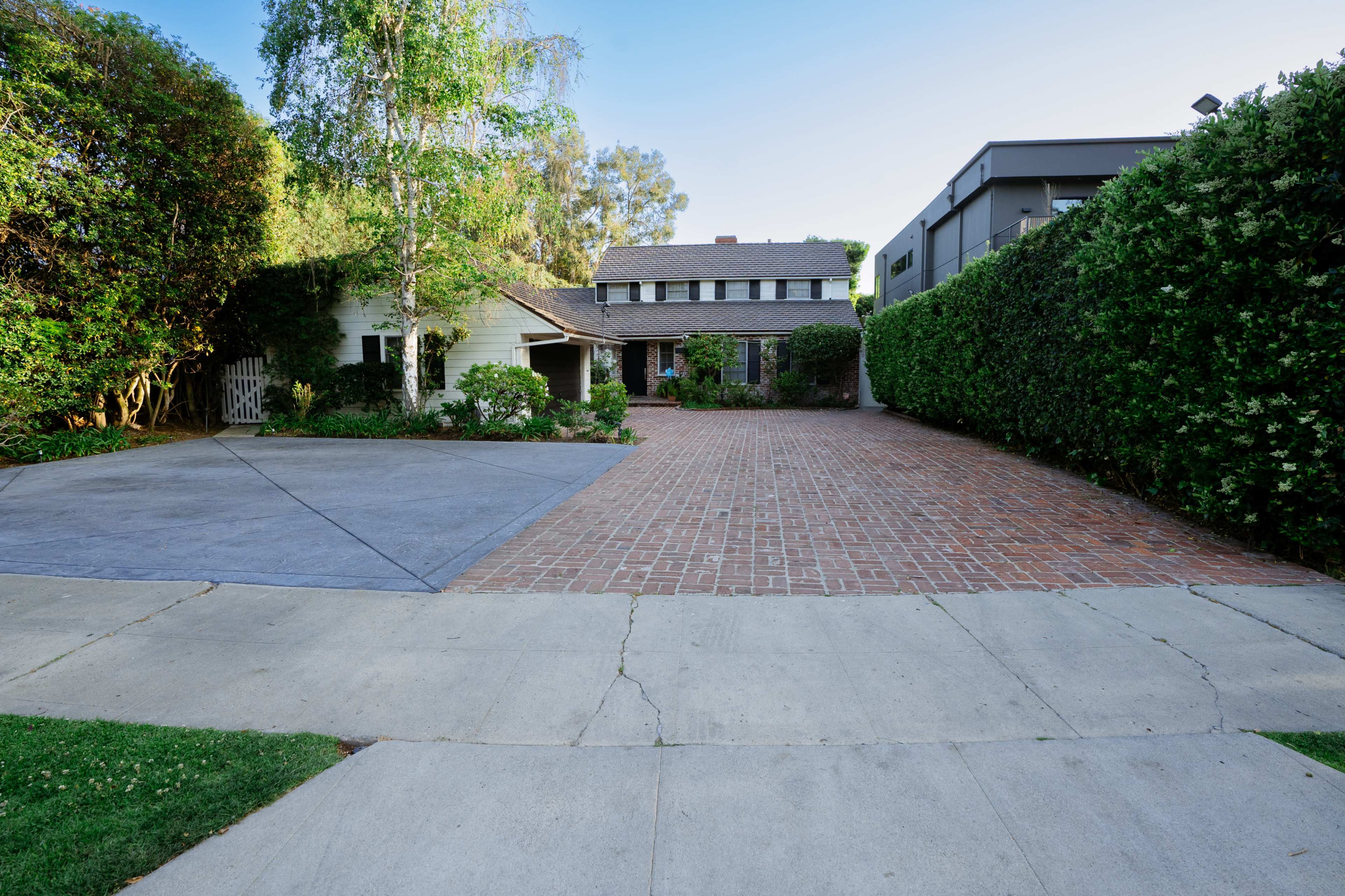 The image shows a residential property with a brick driveway and a house set back from the street, surrounded by manicured shrubs and trees.