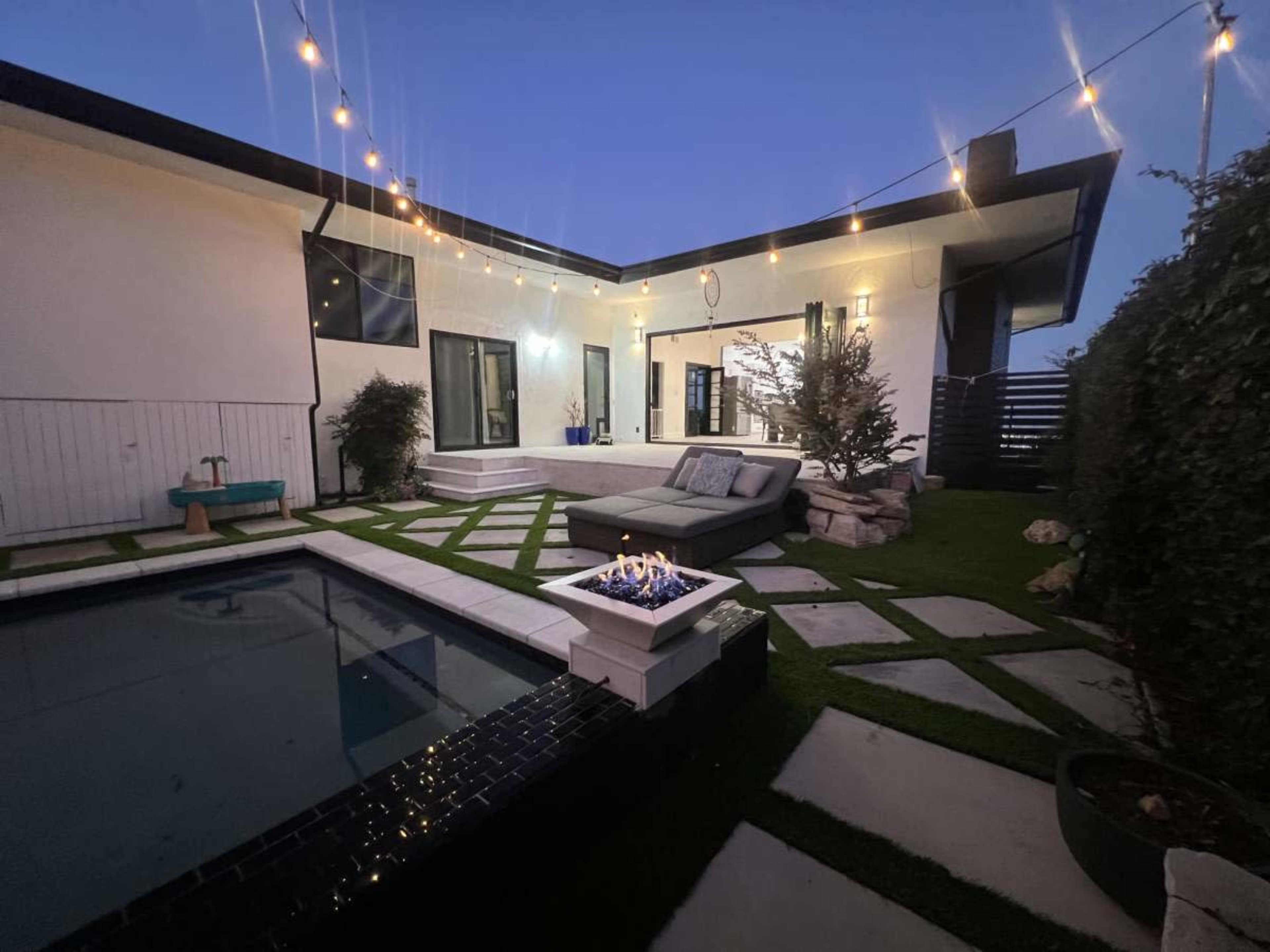The image shows a modern outdoor space featuring a pool, a fire pit with seating, and string lights overhead, surrounded by greenery and stone pathways.