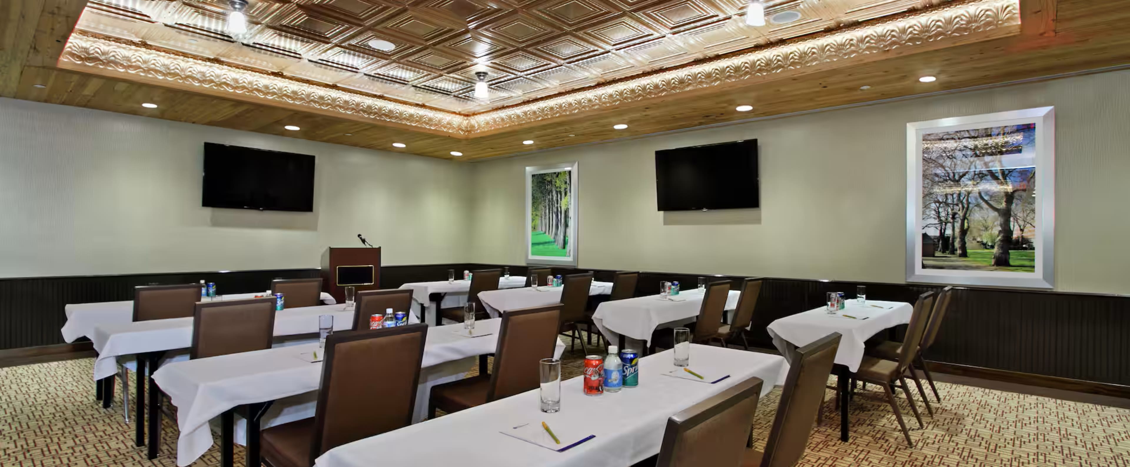 The image depicts a conference room set up with rows of tables covered in white tablecloths, accompanied by chairs, and two large television screens mounted on the walls.