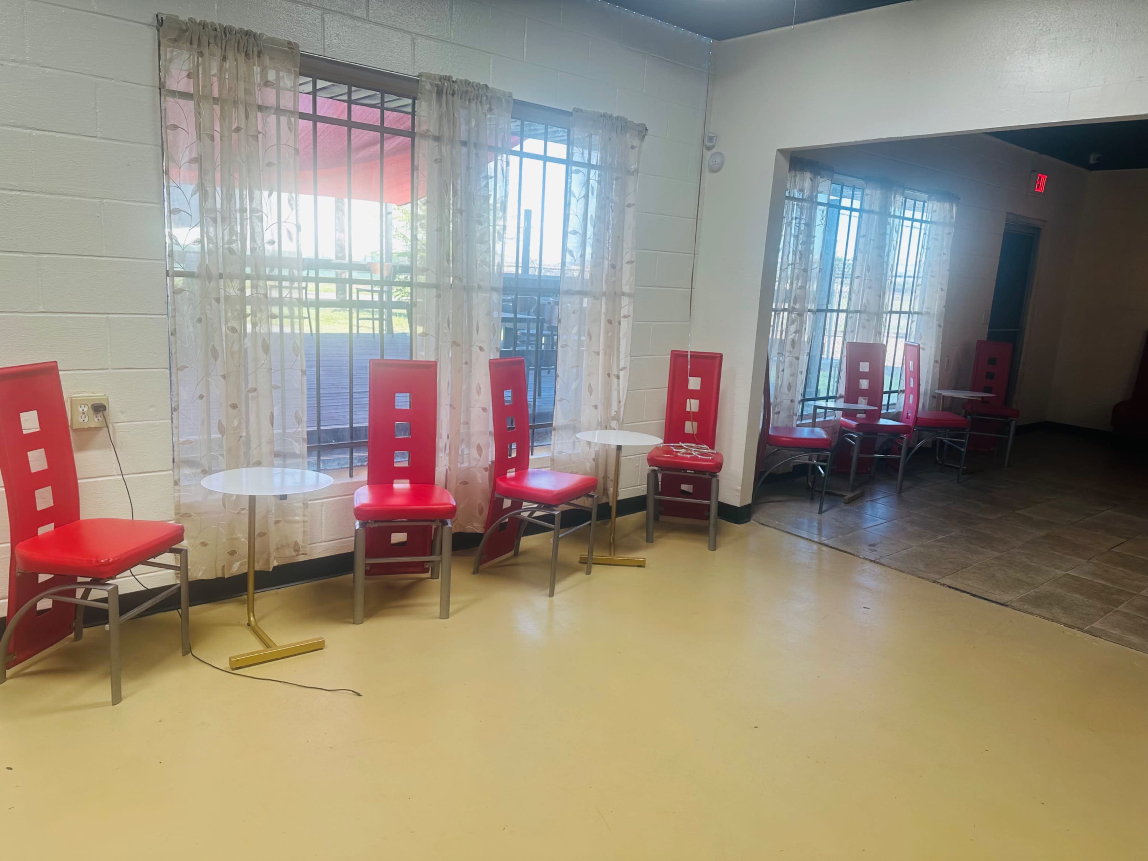 A row of red chairs and small tables is positioned near two large windows with sheer curtains in a brightly lit room.