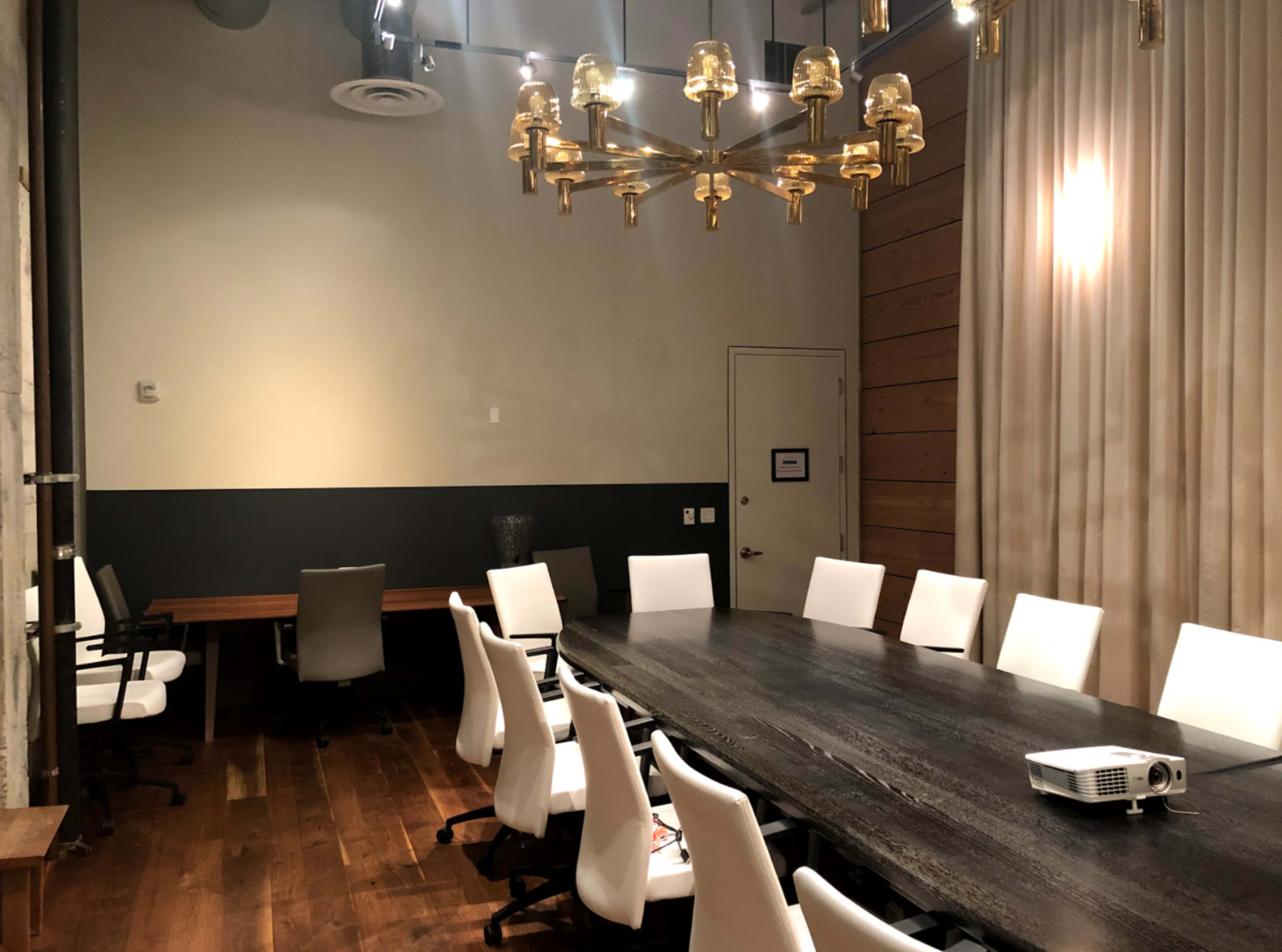 A large conference room features a wooden table surrounded by white chairs, with a projector mounted on the side and a modern lighting fixture overhead.