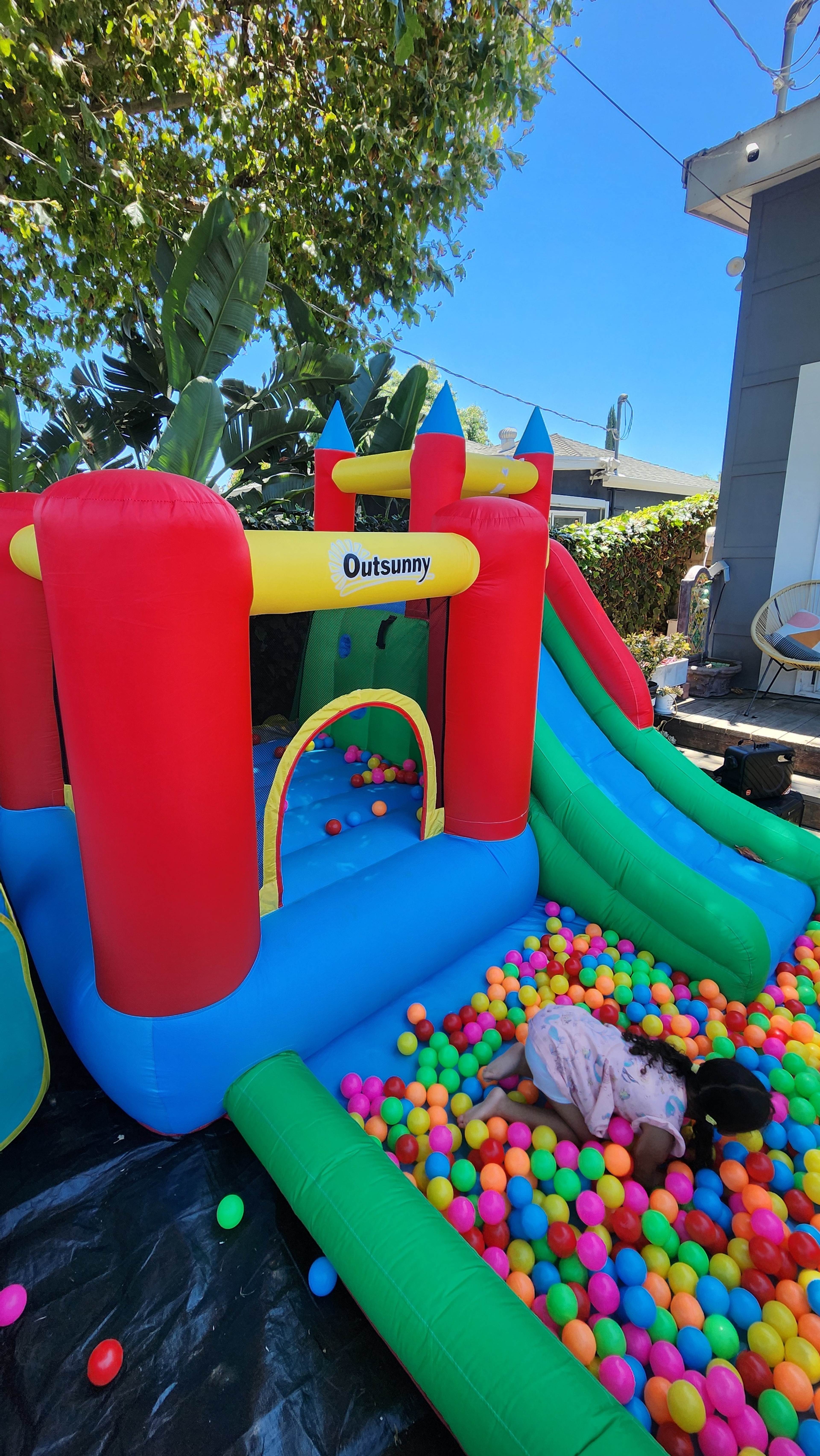 A colorful inflatable bouncy castle with a slide sits in a backyard, while a child plays in a ball pit nearby.