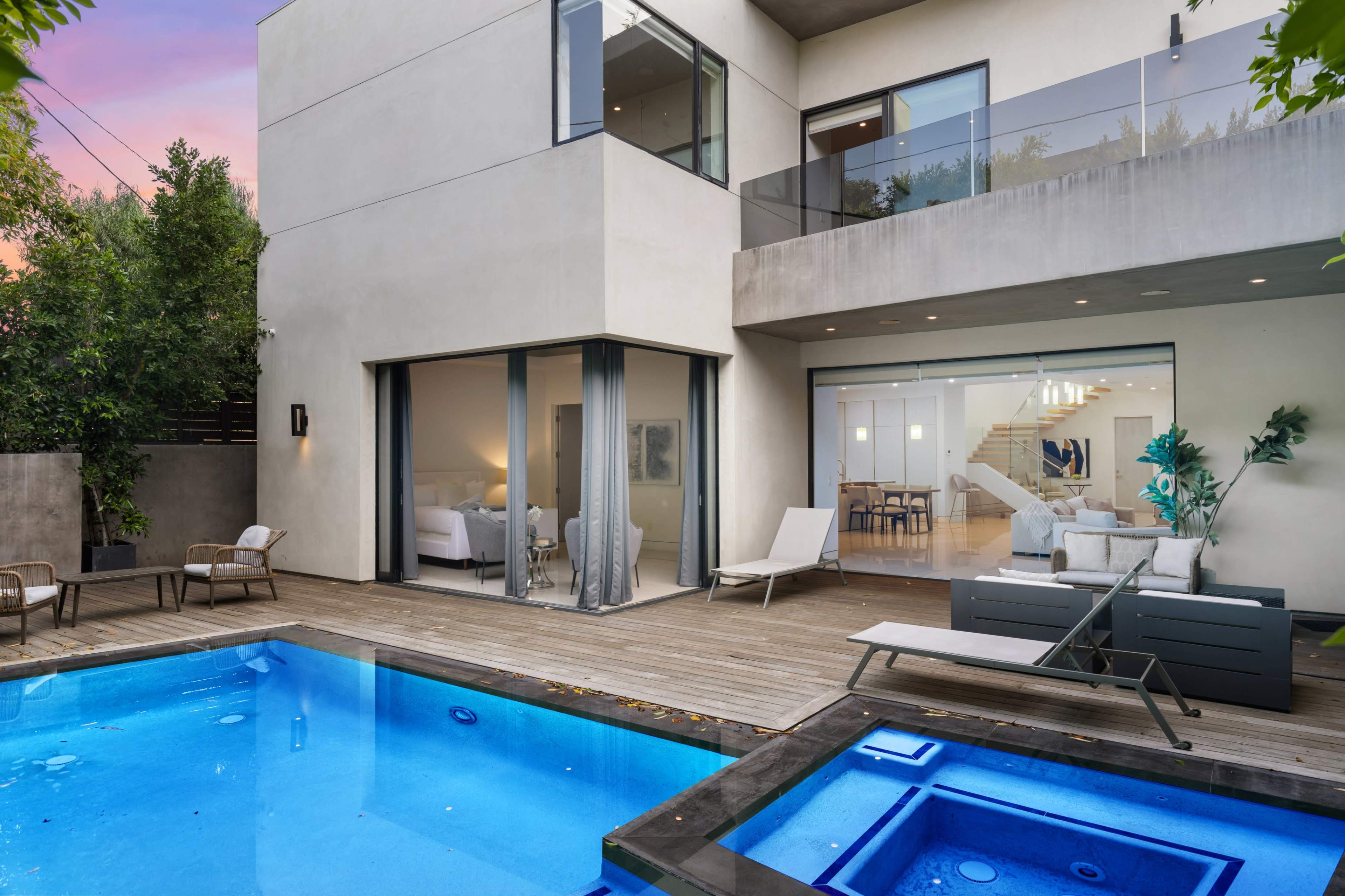 A modern home with a wooden deck surrounds a blue swimming pool, and large glass doors open to an outdoor living area and a staircase leading to the interior.