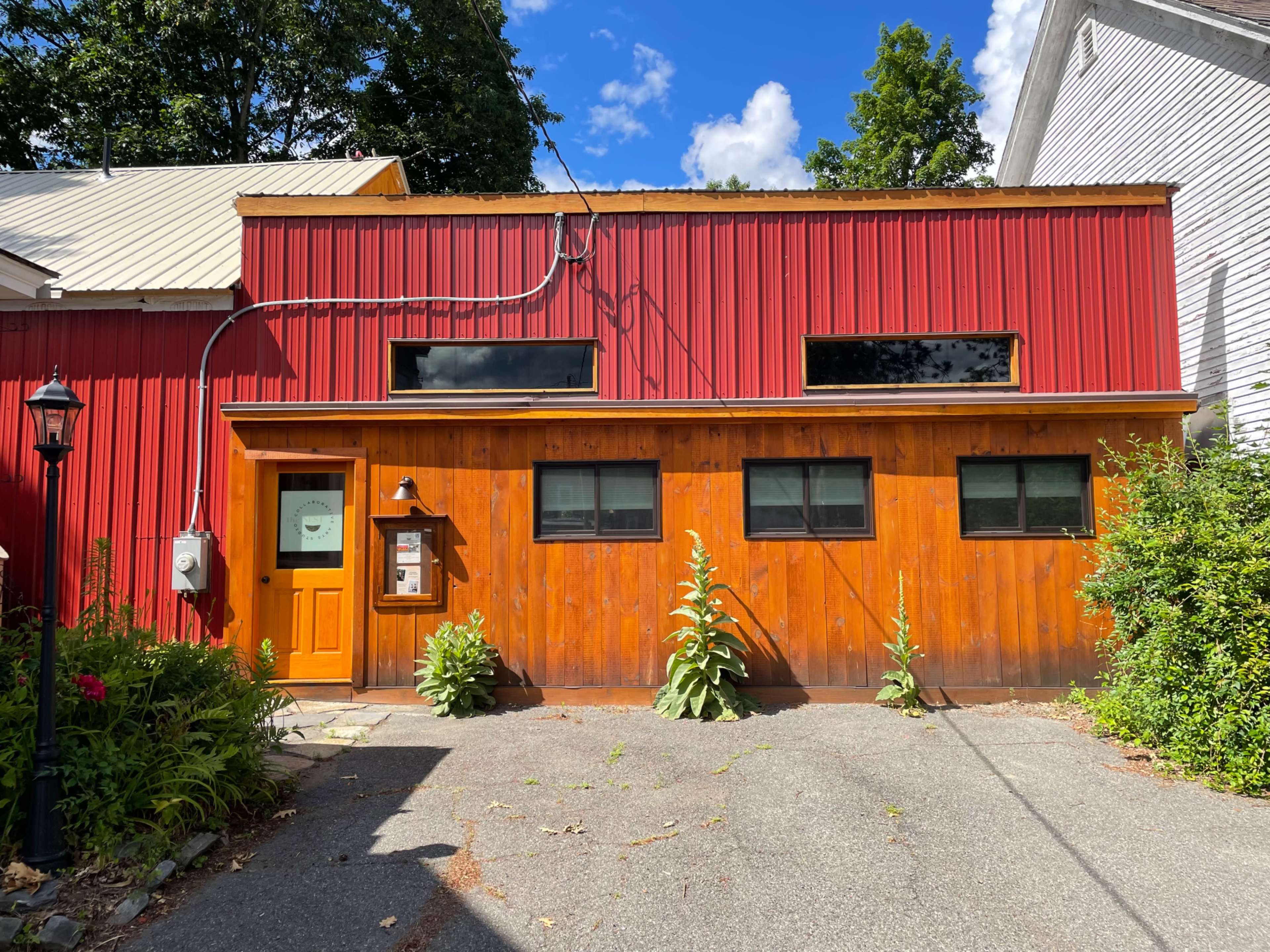 A red and wooden building with multiple windows, situated along a paved path and surrounded by greenery.