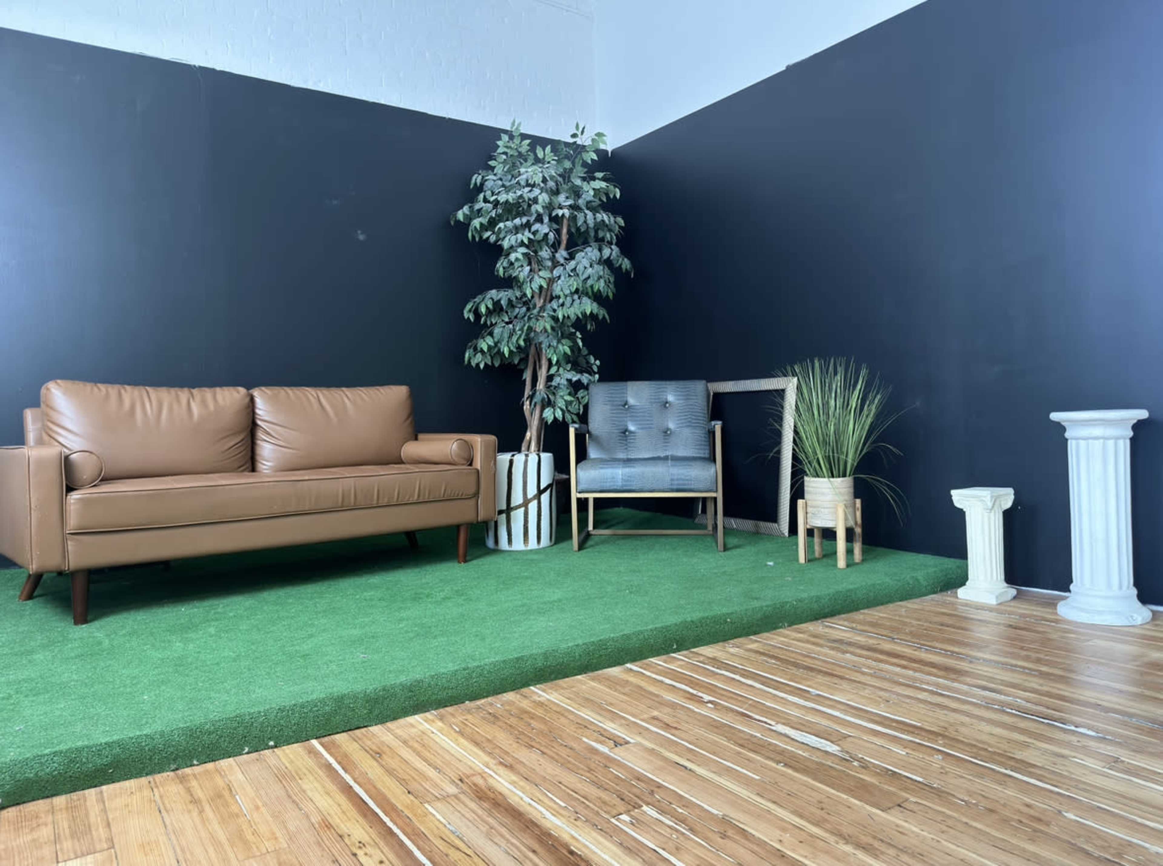 The image shows a small interior space with a brown couch, a gray chair, decorative plants, and a wooden floor.