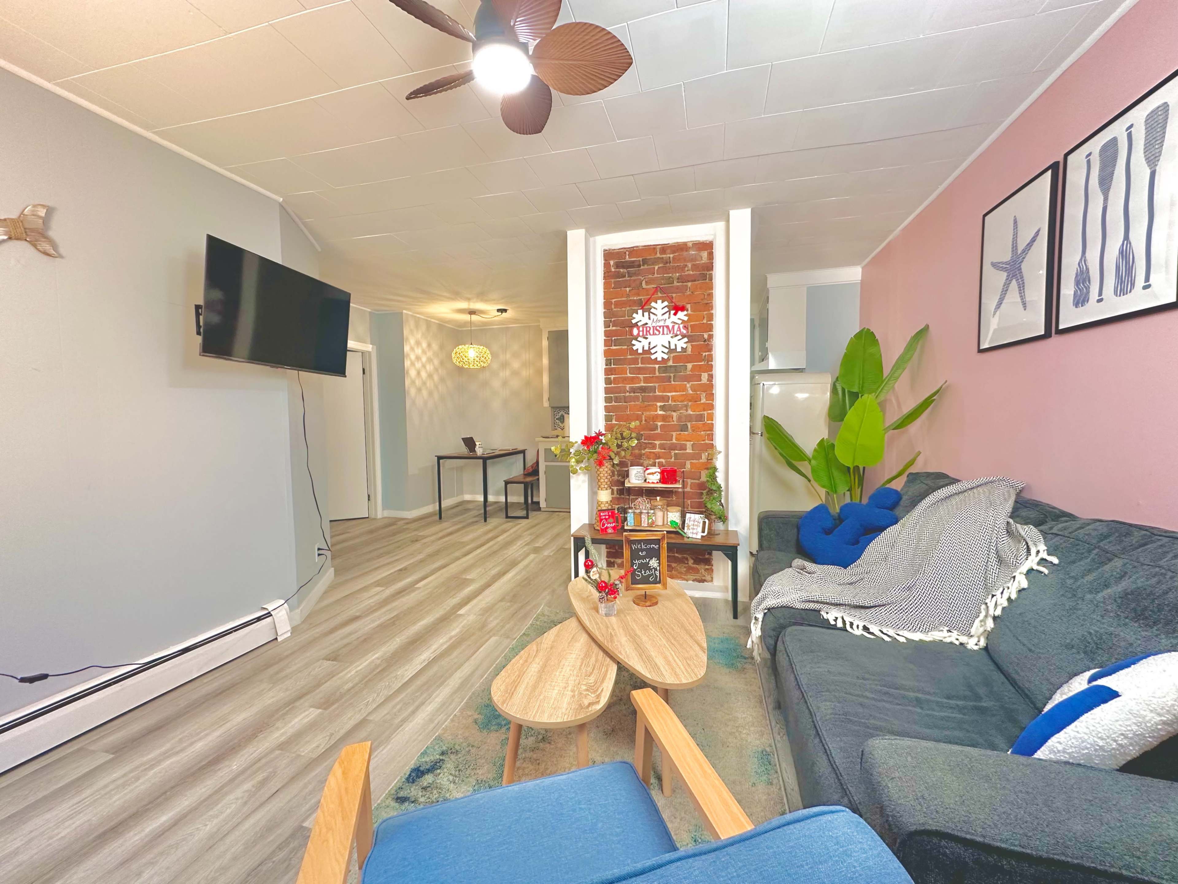 The image shows a cozy living room with a blue couch, wooden coffee tables, a television mounted on the wall, and decorative elements like a plant and a brick accent wall.