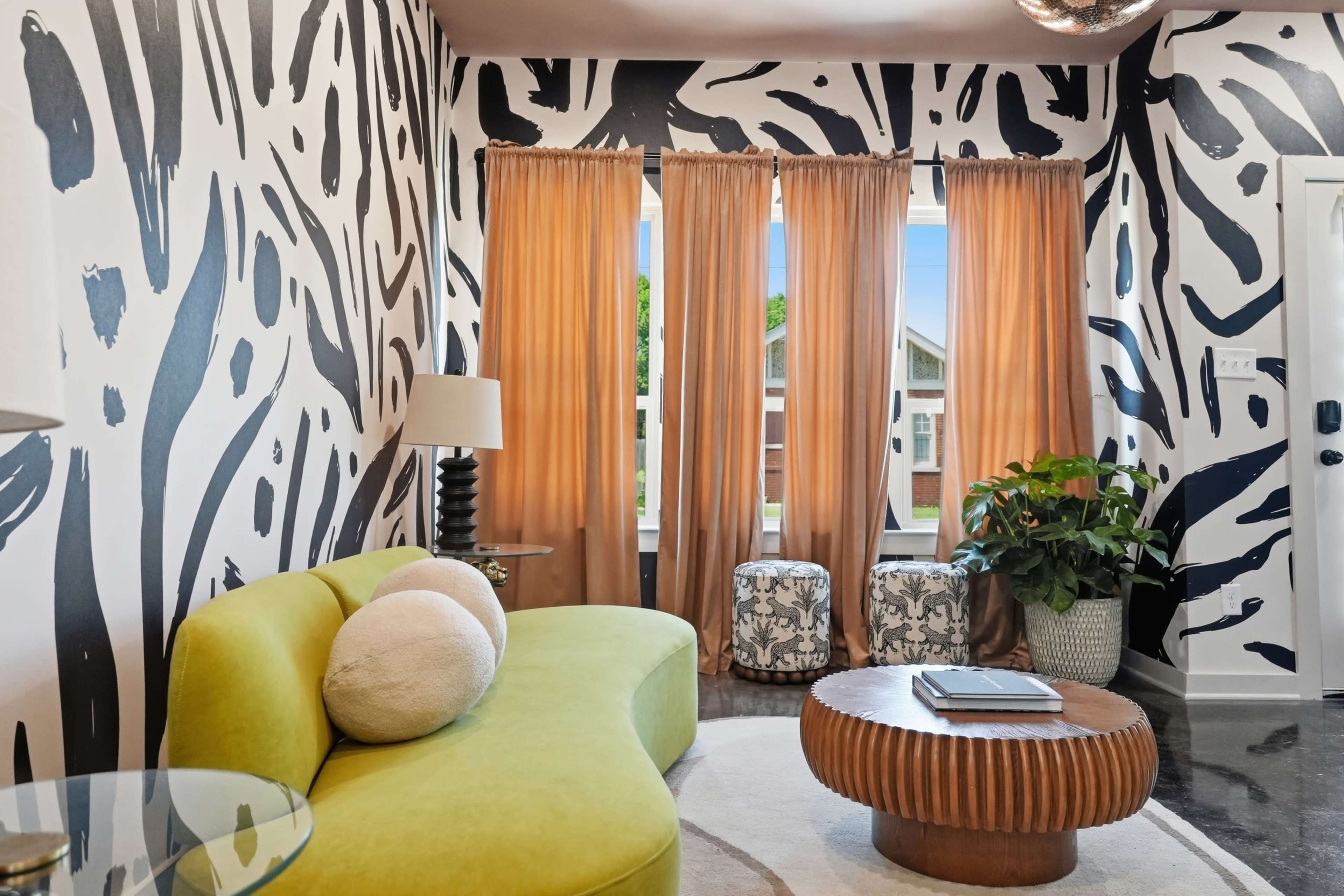 The image shows a living room with bold black-and-white patterned walls, a curved green sofa, a round wooden coffee table, and large windows covered with light curtains.