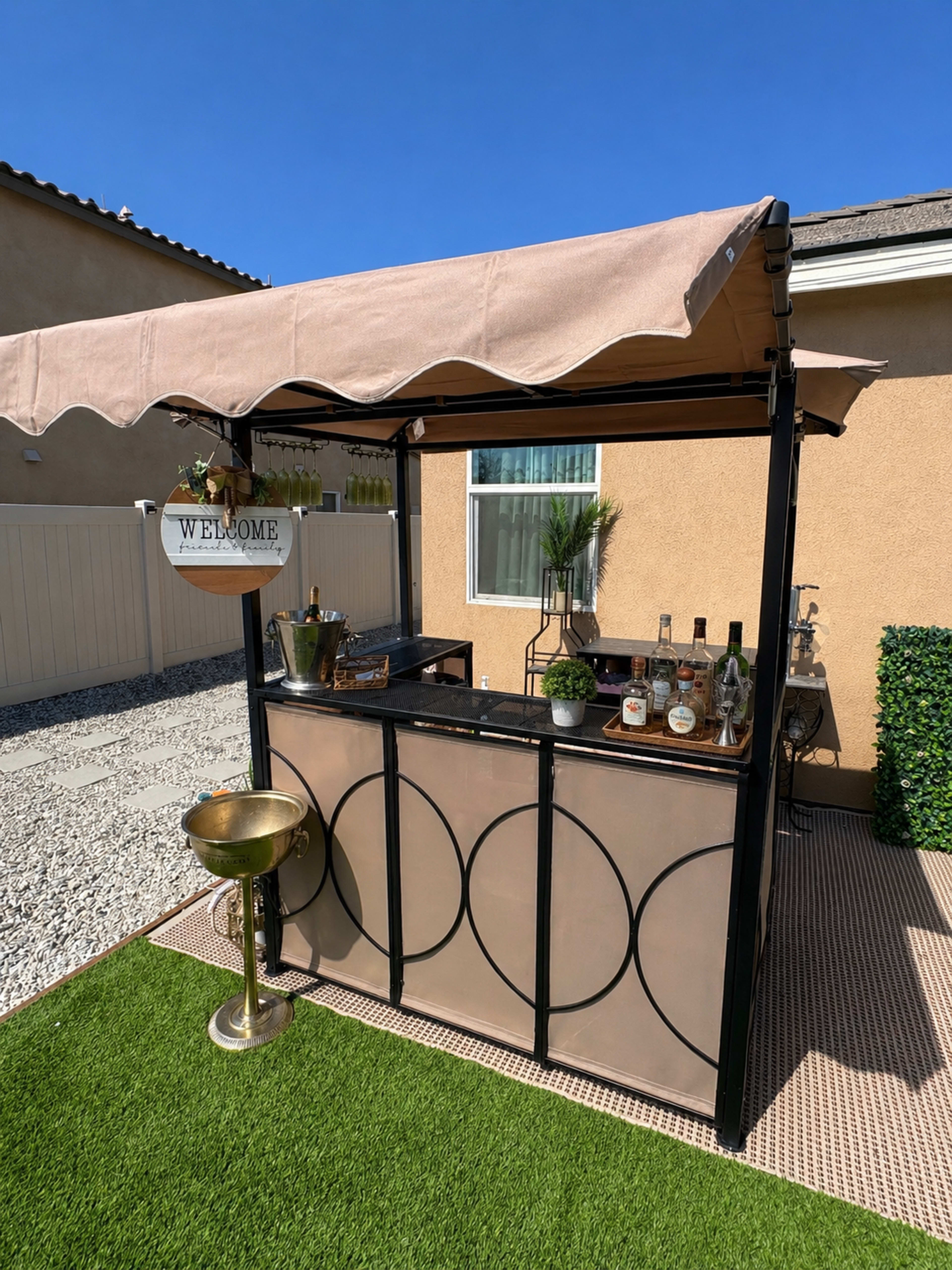 A garden bar with a tan canopy displays various bottles and accessories, set against a backdrop of a beige wall and greenery under a clear blue sky.