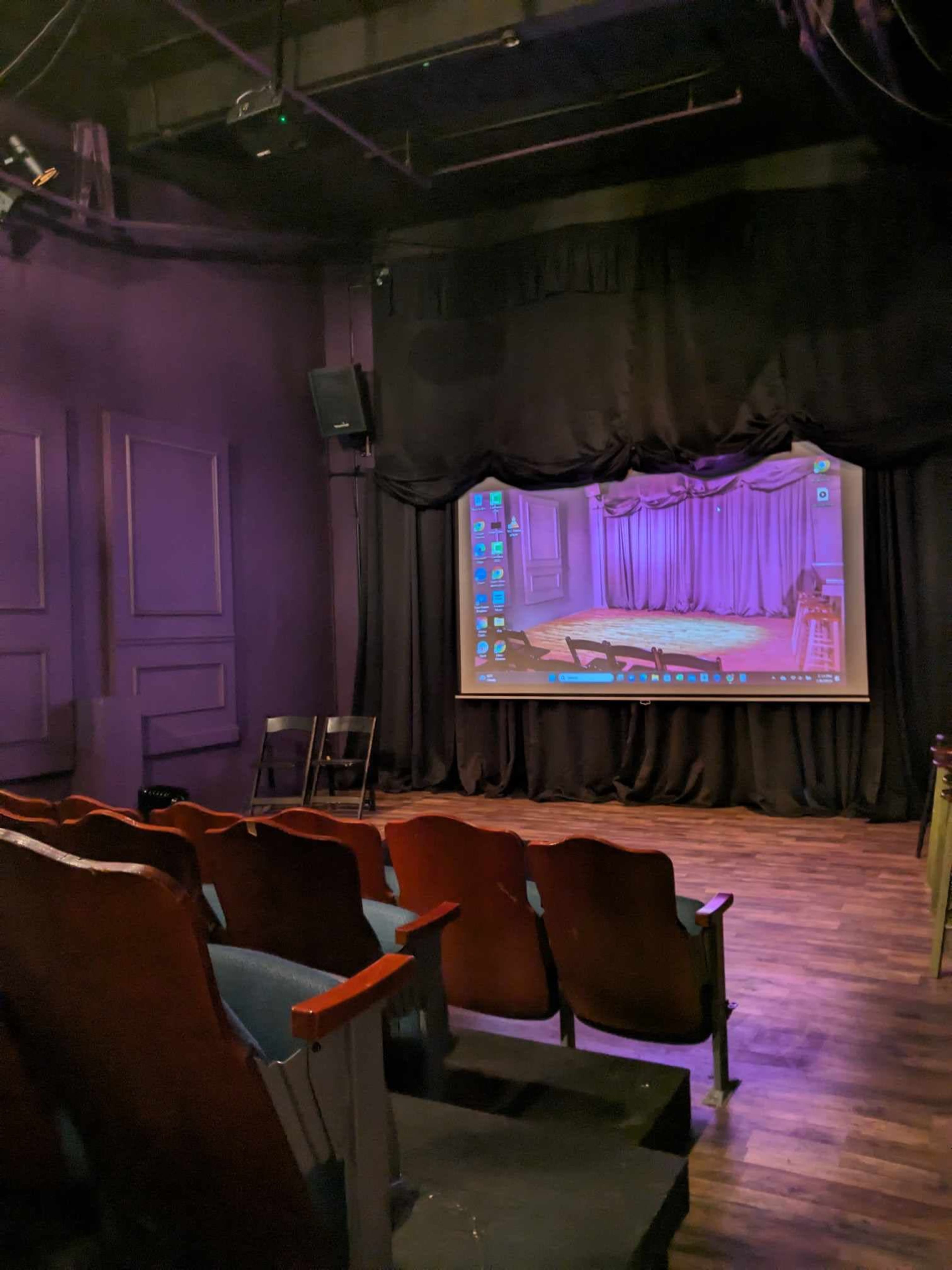 An empty small theater with wooden seats facing a screen and a stage draped in purple curtains.