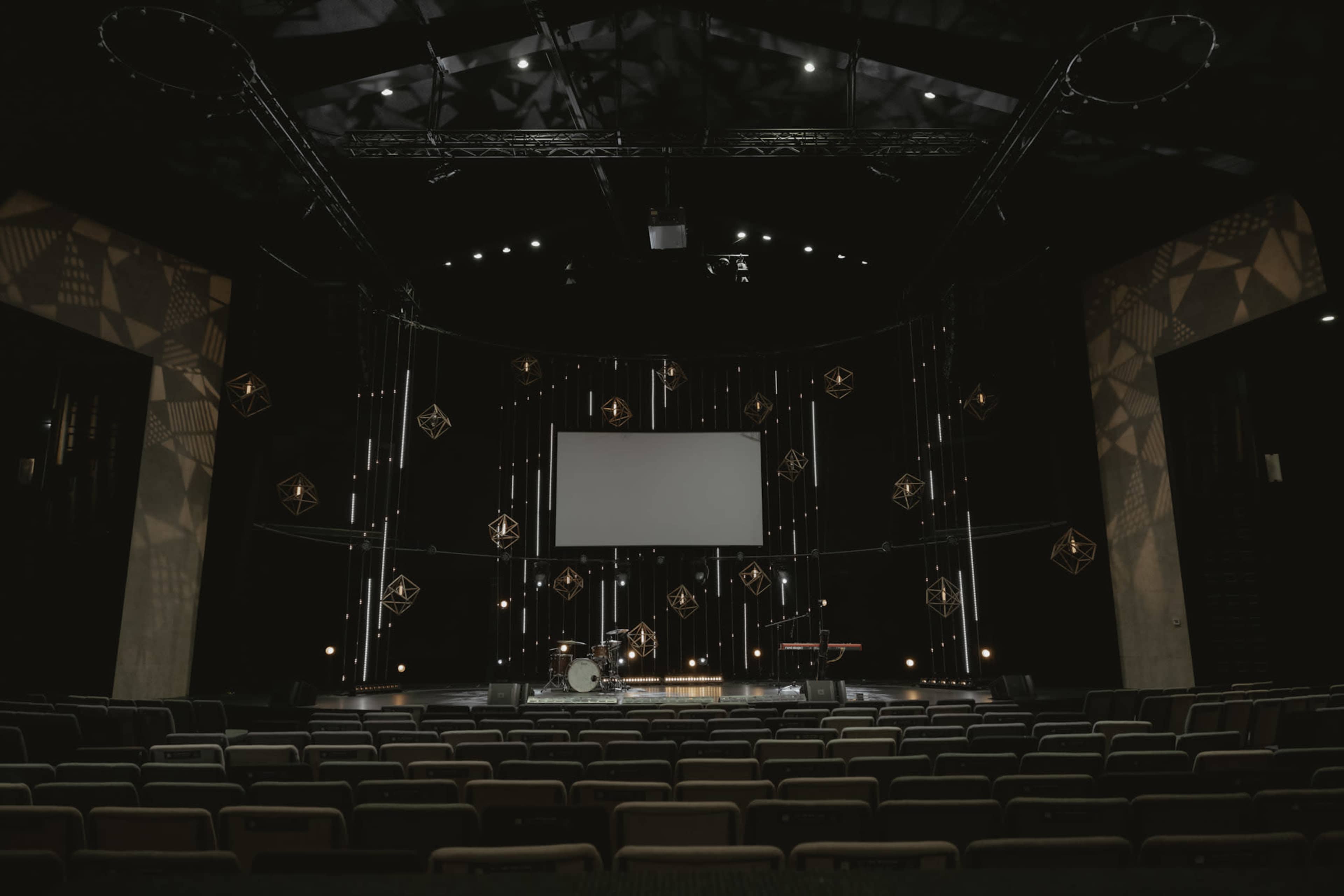 A stage set up for a performance with a blank screen, musical instruments, and lighting in a darkened theater filled with empty seats.