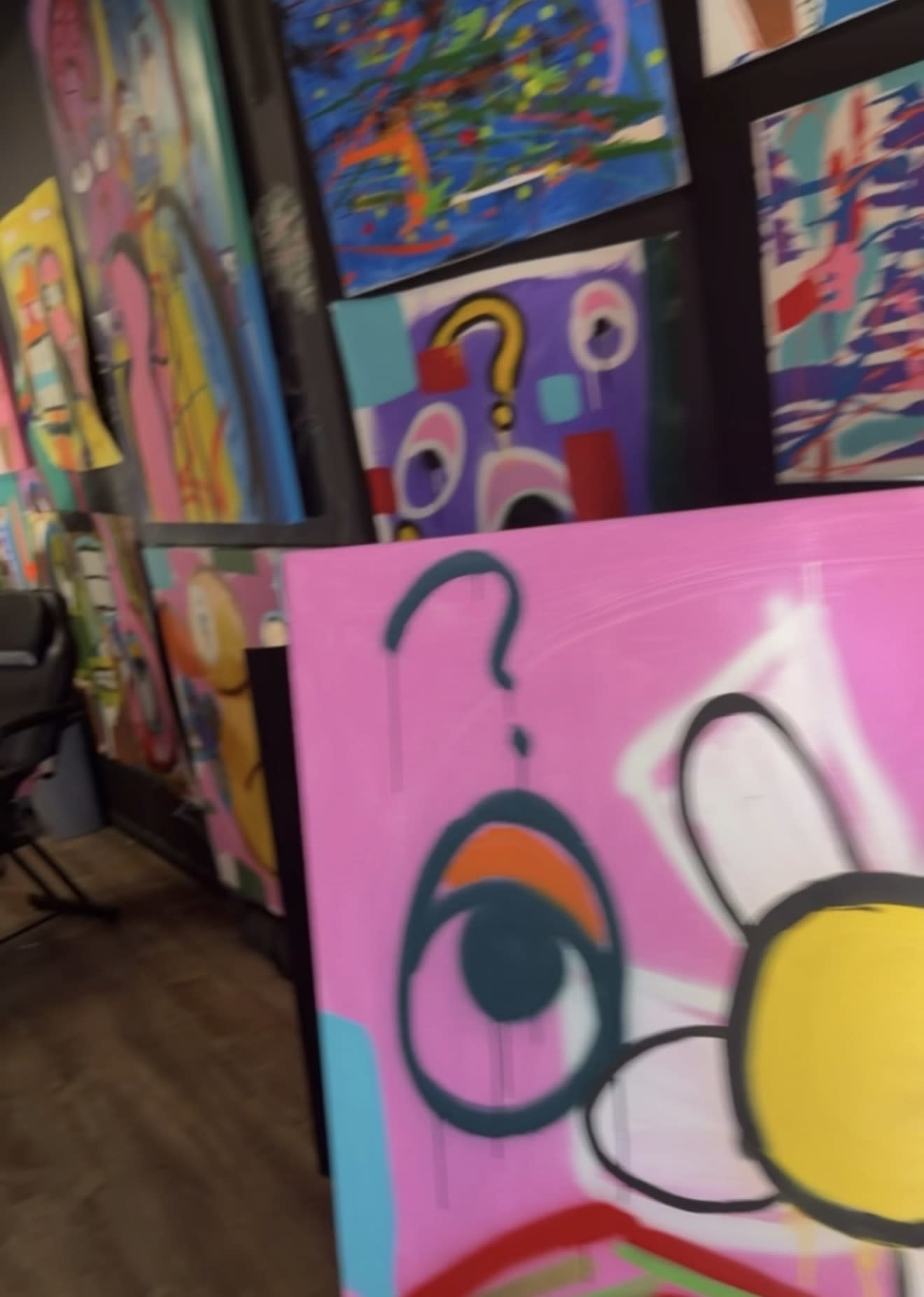 The image shows a colorful art gallery featuring vibrant abstract paintings on the walls, with a focus on a large pink canvas displaying a stylized eye and a daisy.