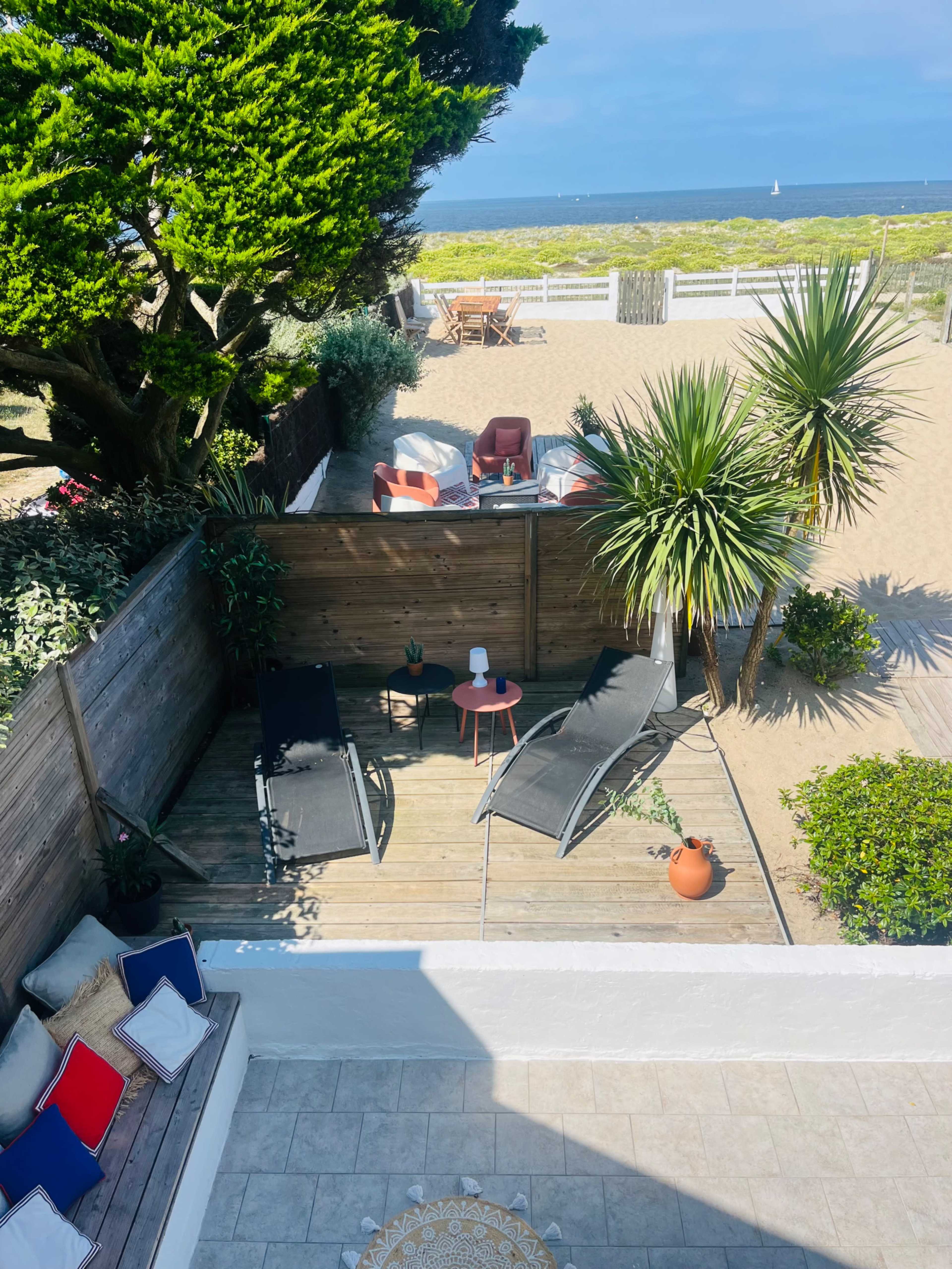 The image shows a sunny outdoor space with two lounge chairs and a small table surrounded by tropical plants and a sandy beach in the background.