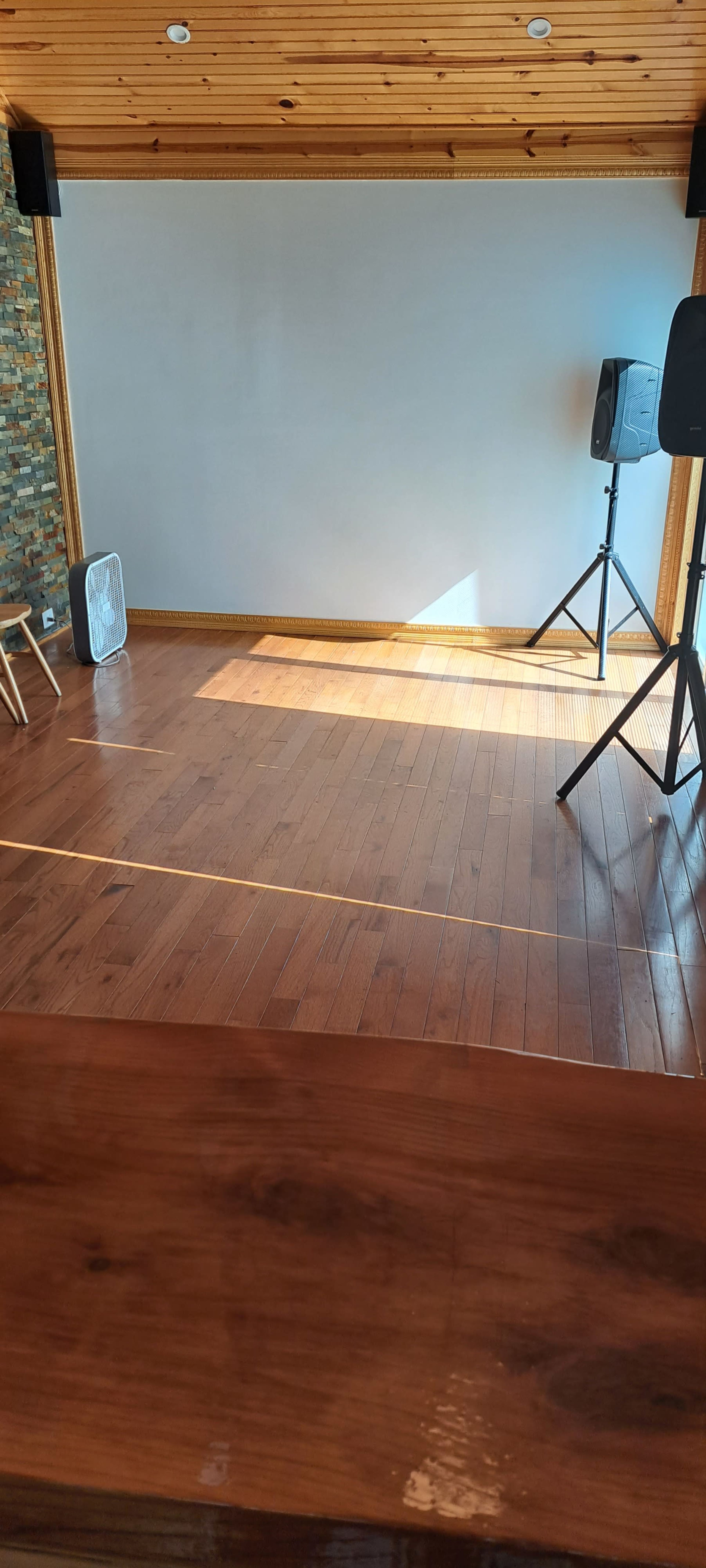 A wooden floor space with a fan and two speaker stands, set against a stone wall and a blank area for projection.