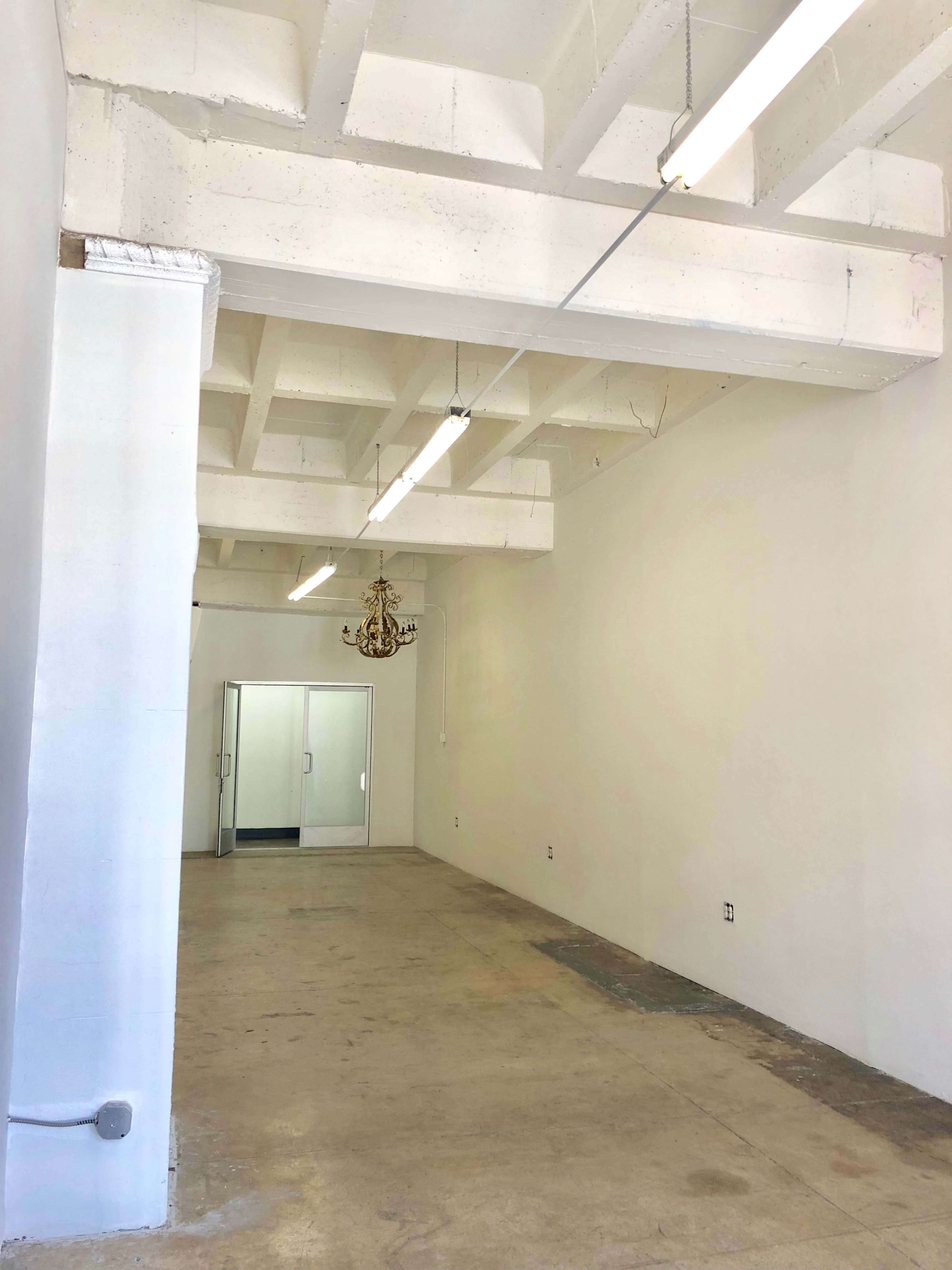 The image shows a spacious, empty room with white walls, concrete flooring, and a chandelier hanging from the ceiling.