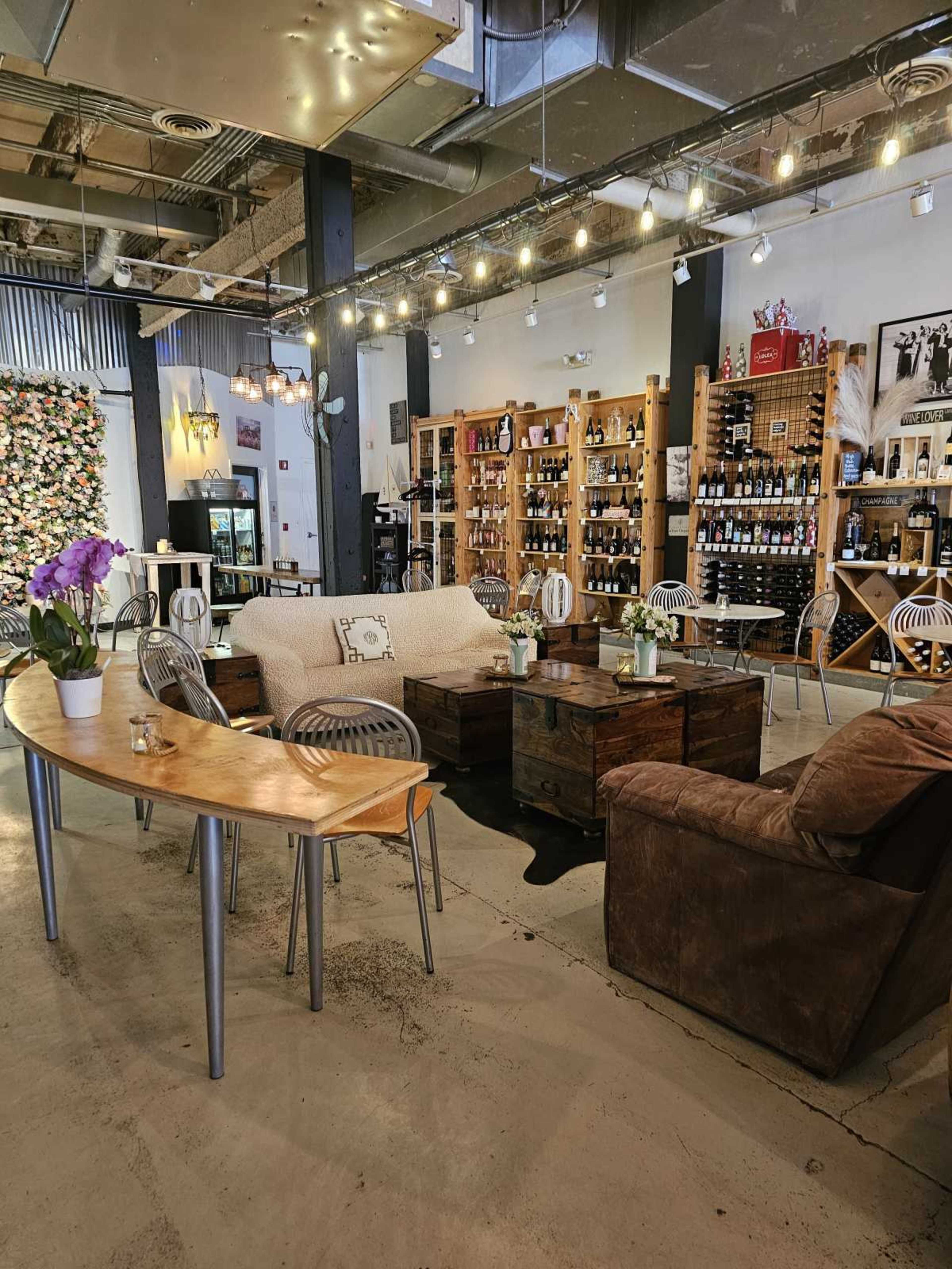 The image shows a cozy seating area in a wine shop, featuring wooden furniture, shelves filled with wine bottles, and decorative lighting.