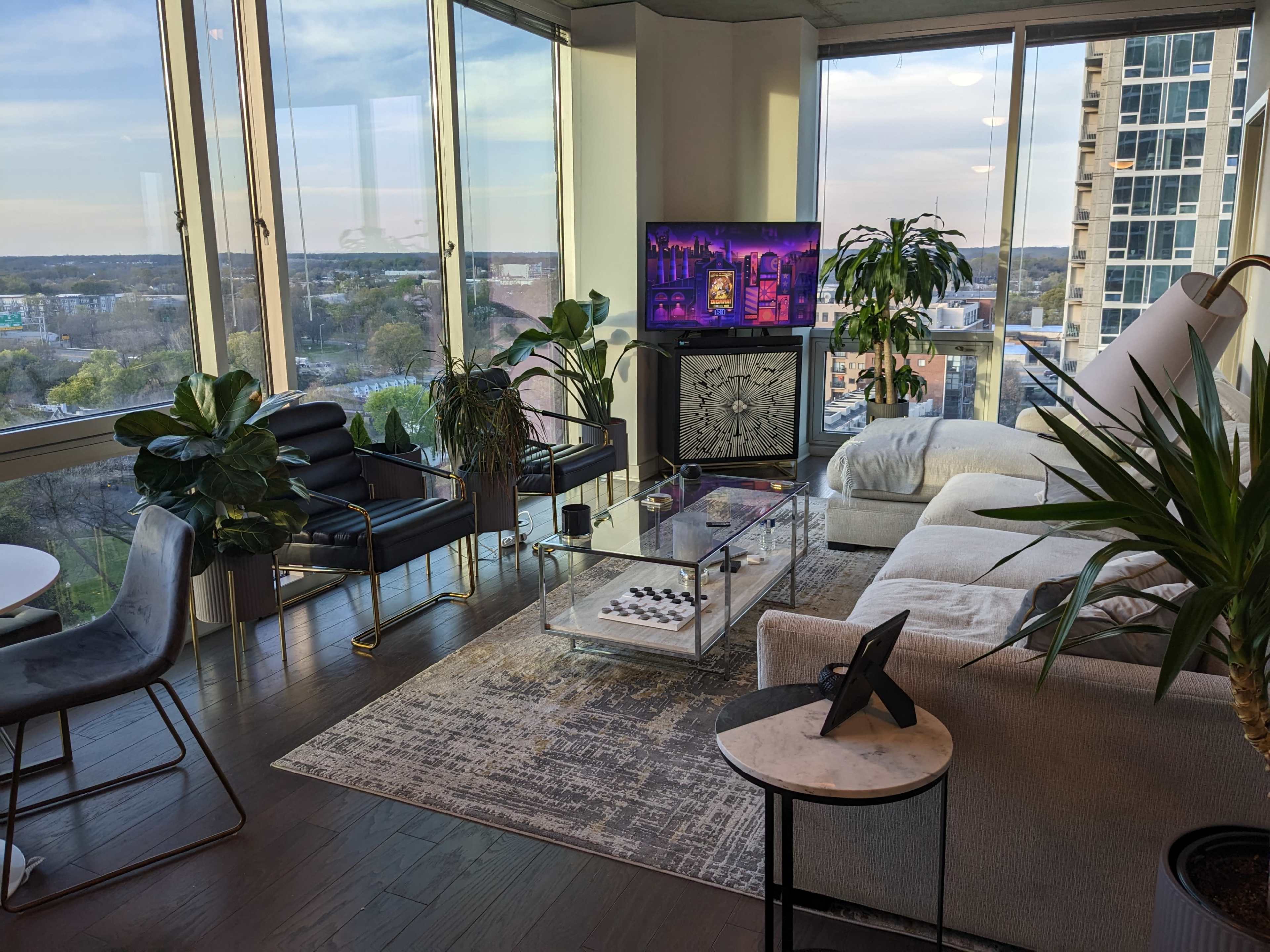 A modern living room with large windows, featuring a television, a coffee table with a chess set, and various indoor plants.
