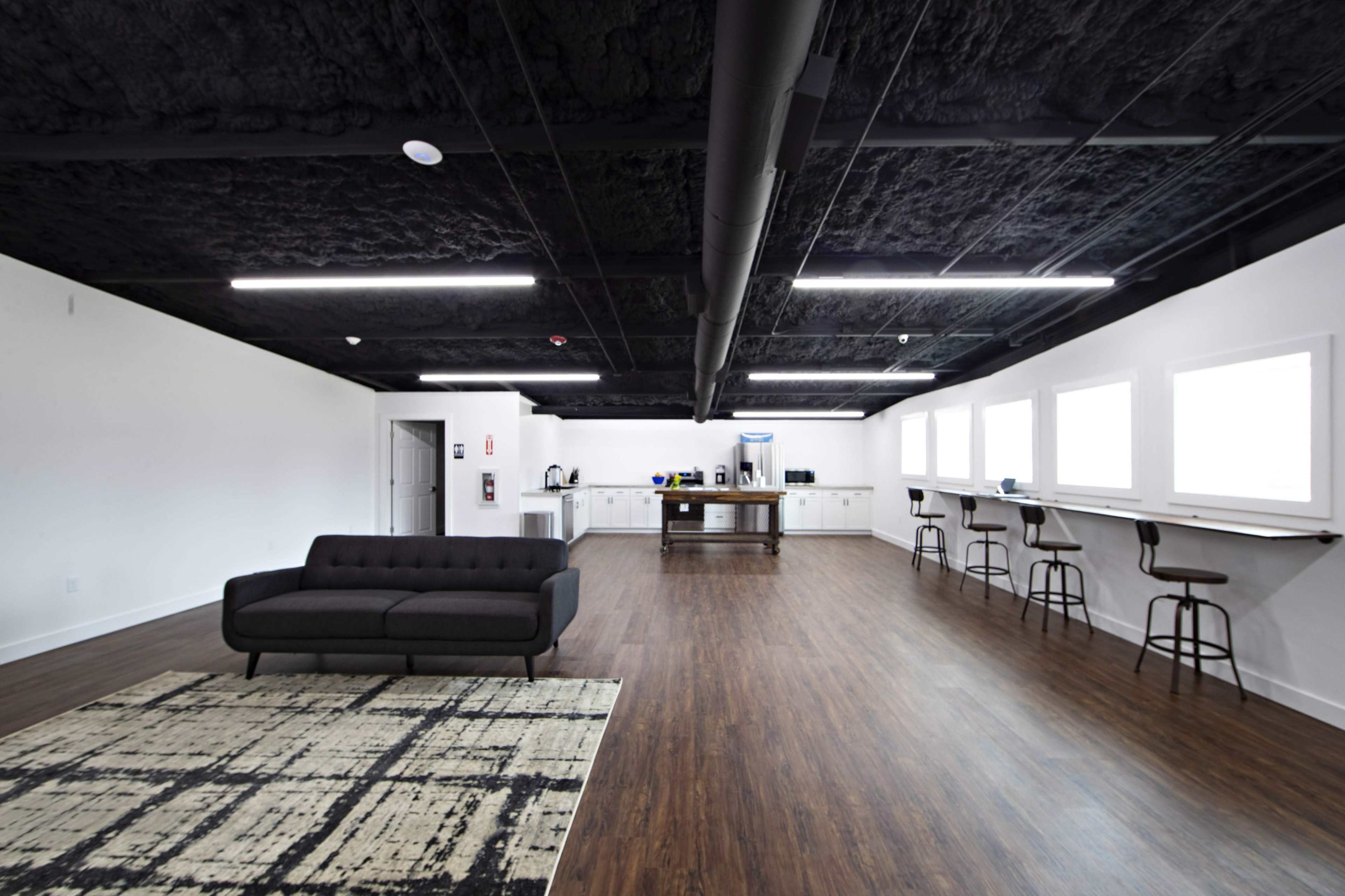 A spacious modern office interior with a black couch, hardwood flooring, a kitchen area, and multiple windows along one wall.