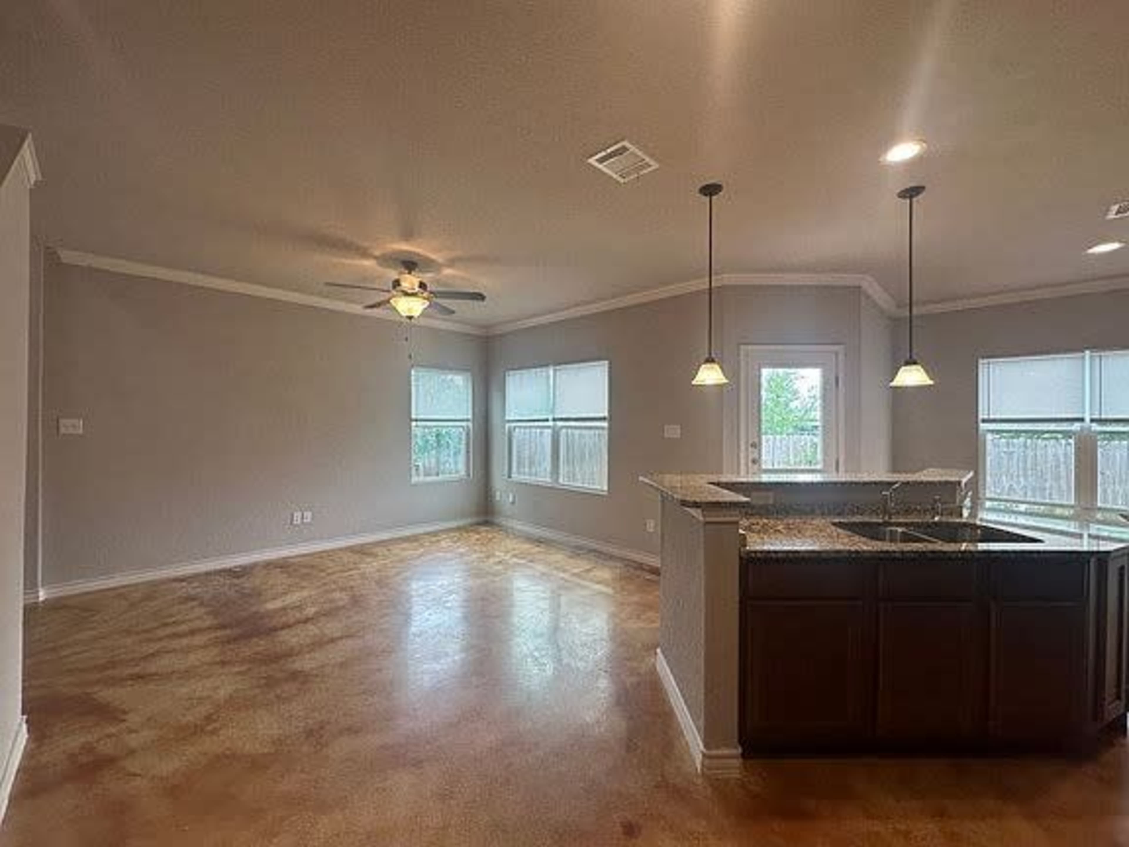 The image shows a spacious living area with a ceiling fan, a small kitchen bar, and several windows providing natural light.