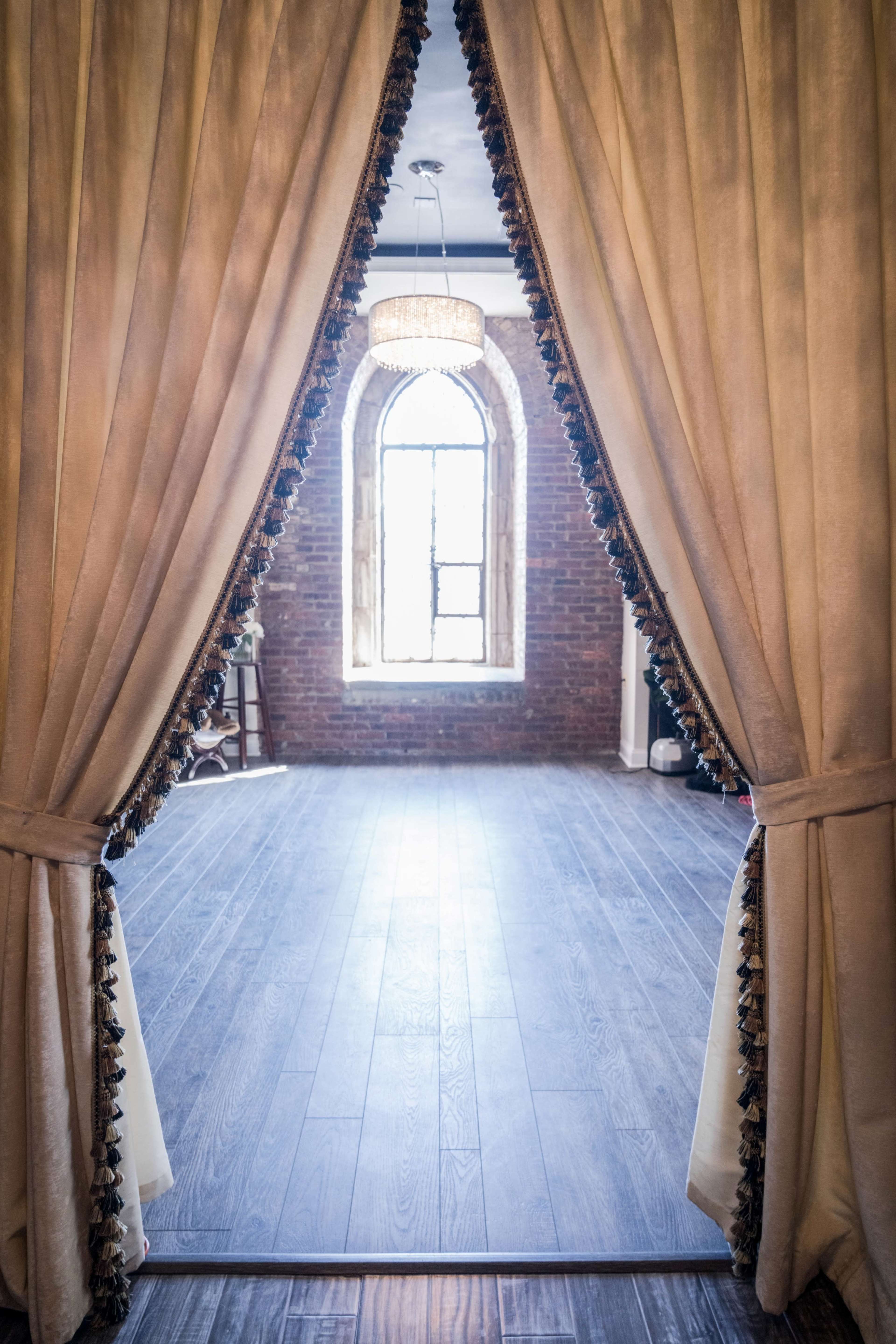 A pair of curtains is parted to reveal a room with a wooden floor and a large arched window in the background.