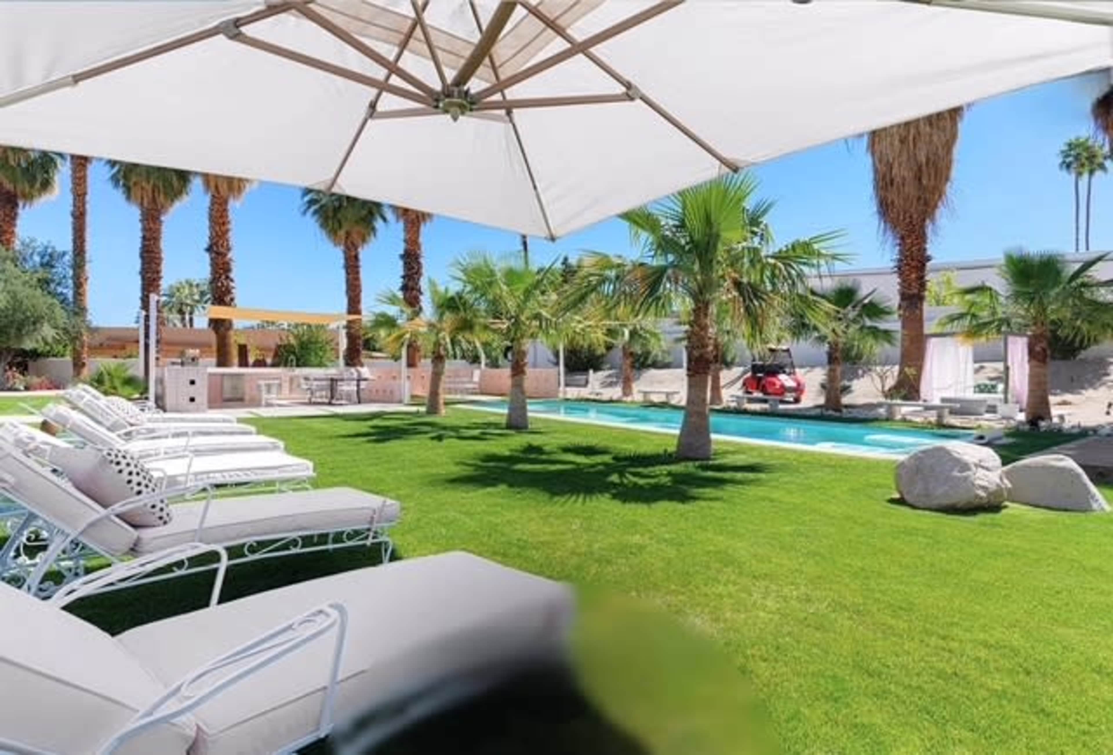 The image shows a lush green lawn with lounge chairs under a large umbrella, palm trees surrounding a swimming pool, and a barbecue area in the background.