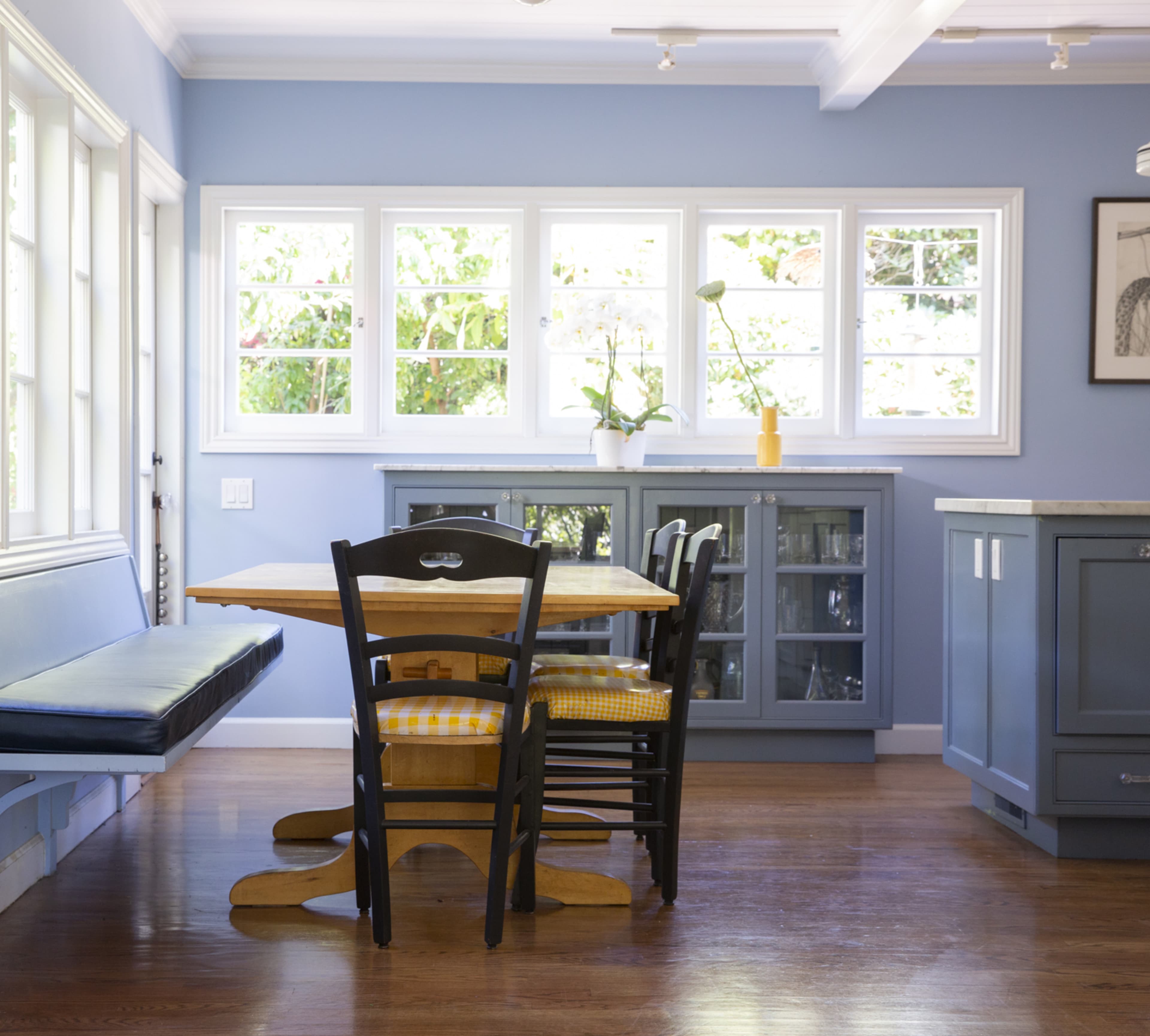 A bright kitchen features a wooden table and black chairs set against blue walls, with large windows letting in natural light.
