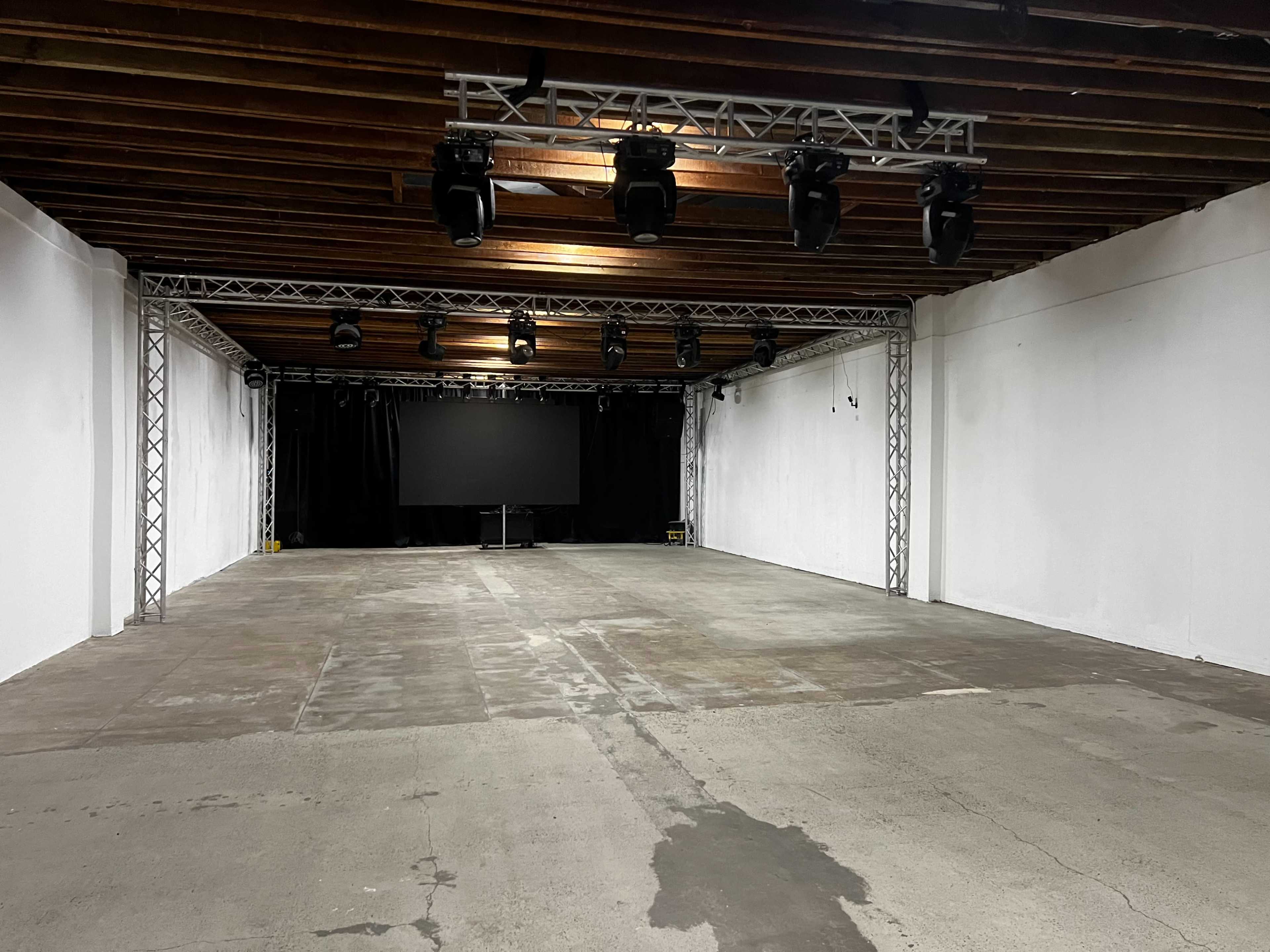 An empty indoor venue with exposed wooden beams, a concrete floor, and stage lighting rigged above a dark backdrop.
