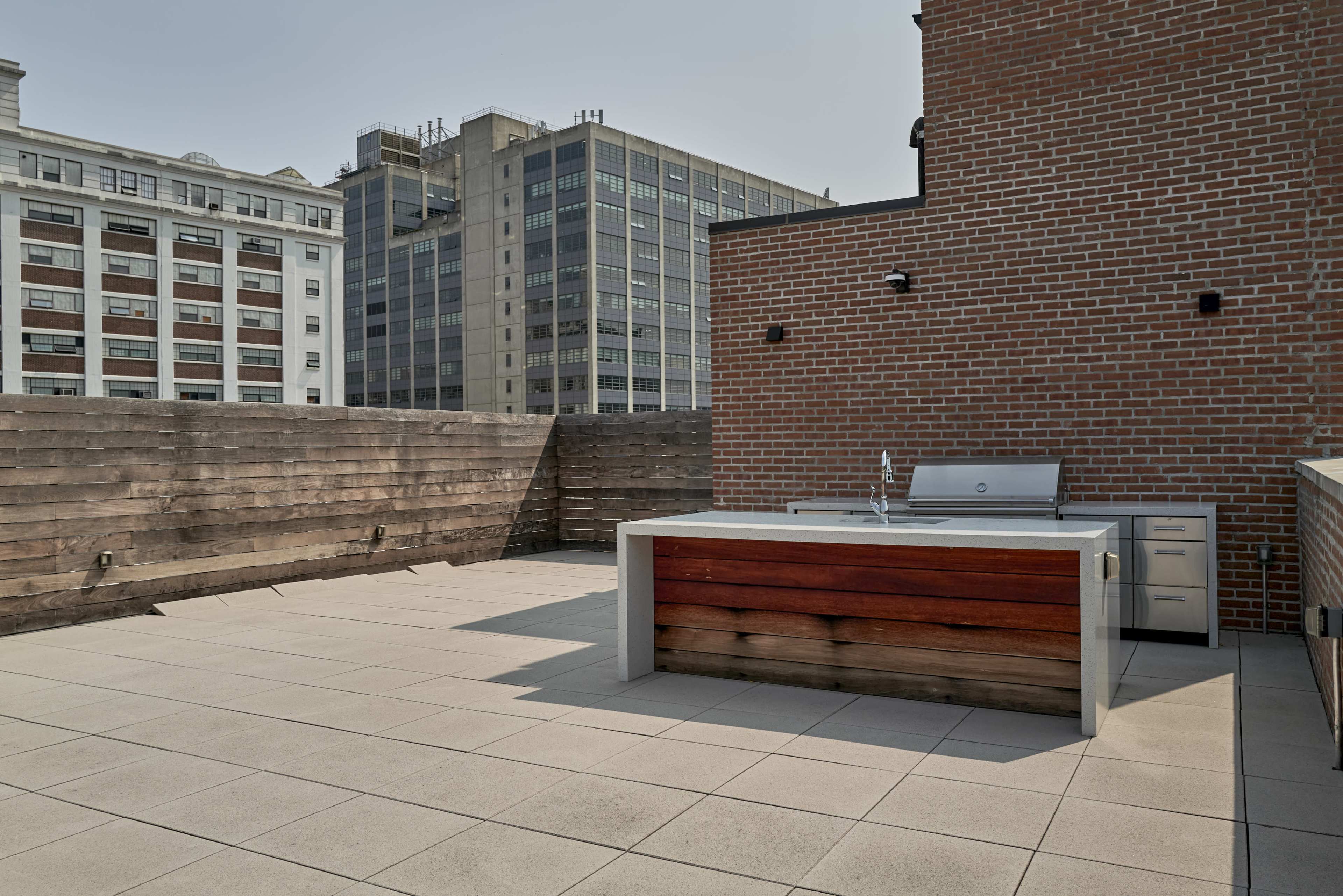 The image shows a rooftop terrace featuring a modern outdoor kitchen with a grill and countertop, surrounded by tall buildings.