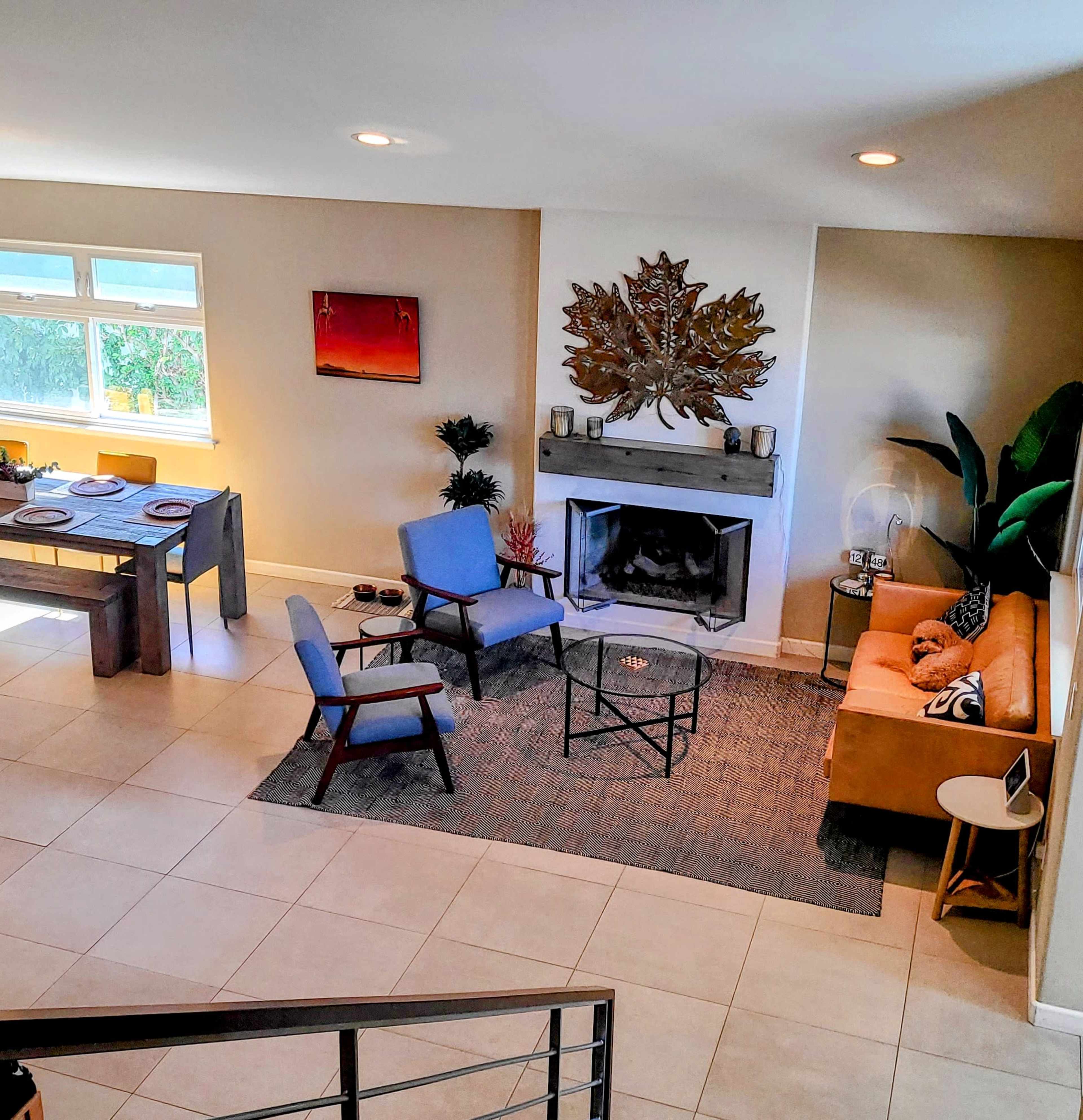 A modern living room with a fireplace, a set of chairs and a sofa, a dining table, and large windows.