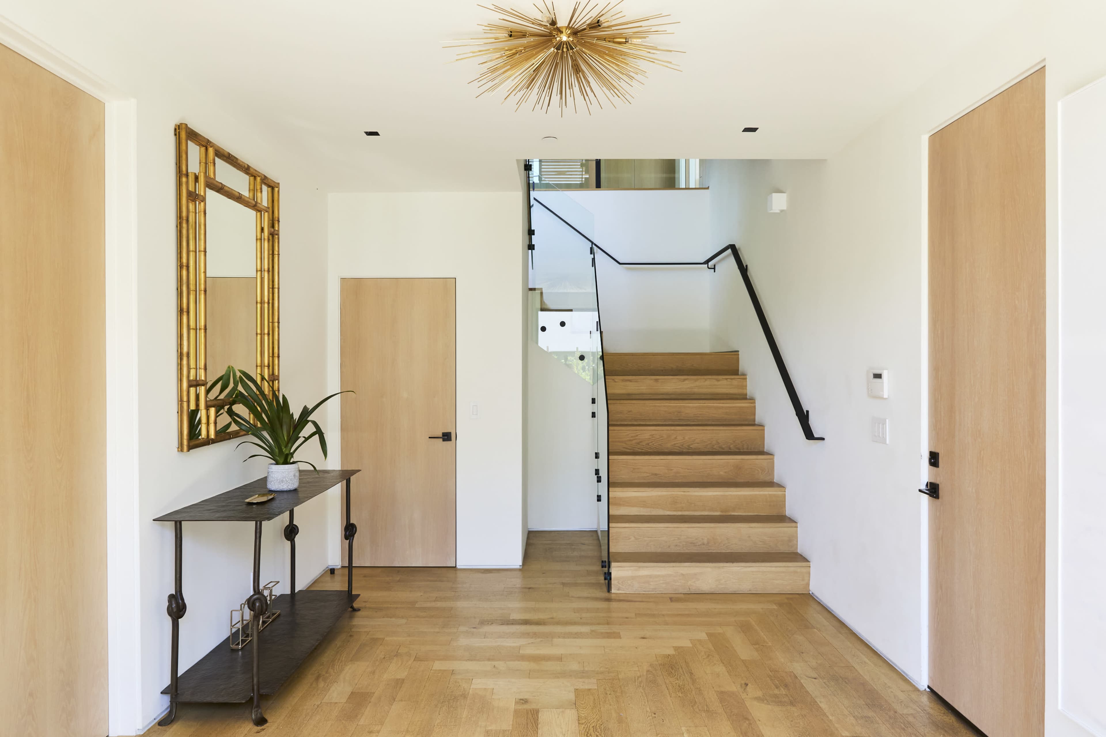 A modern foyer features a wooden staircase, a console table with a plant, and a large decorative mirror on the wall.