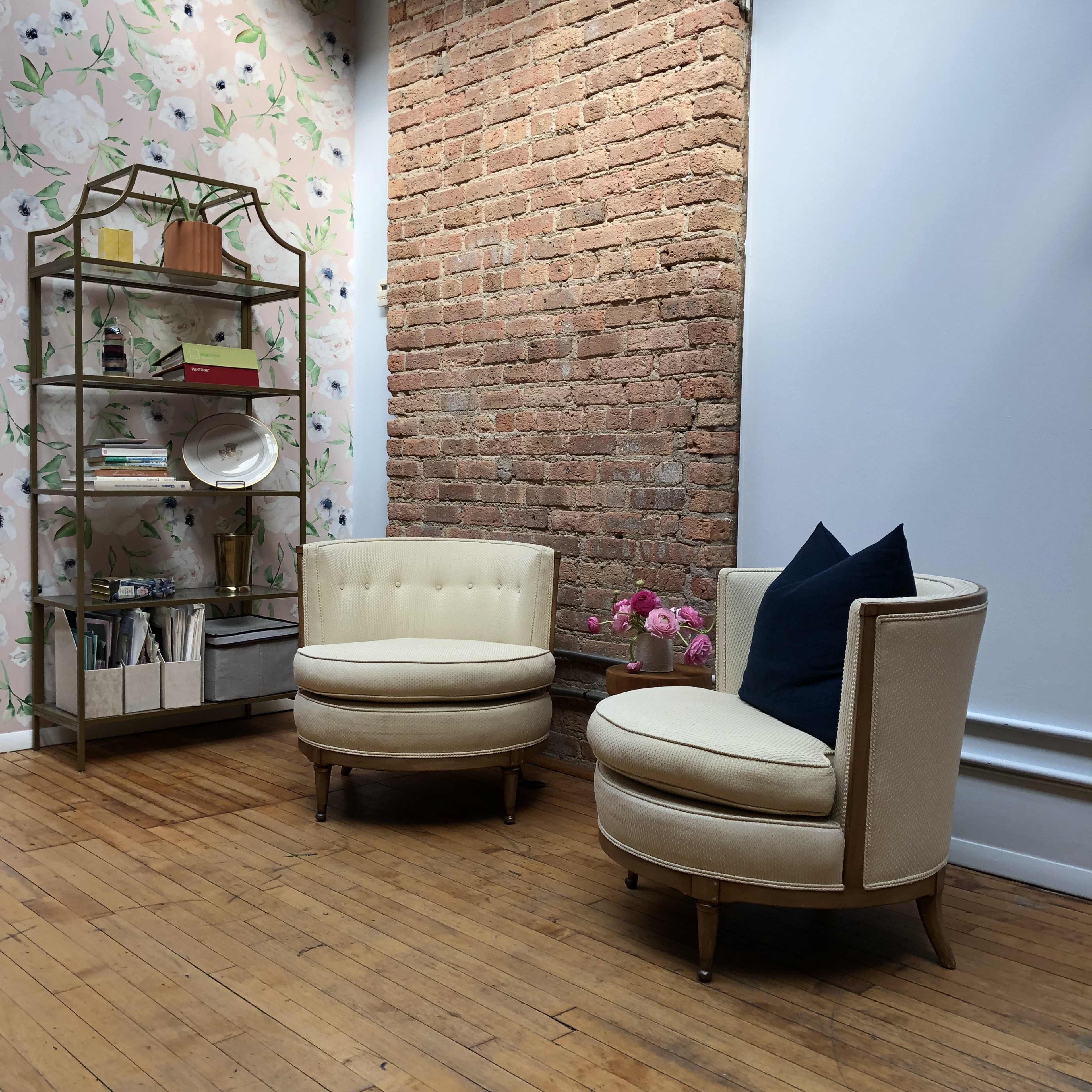 Two beige upholstered armchairs with wooden legs in a room featuring exposed brick walls, a floral wallpaper, and a metallic bookshelf holding decorative items and books.