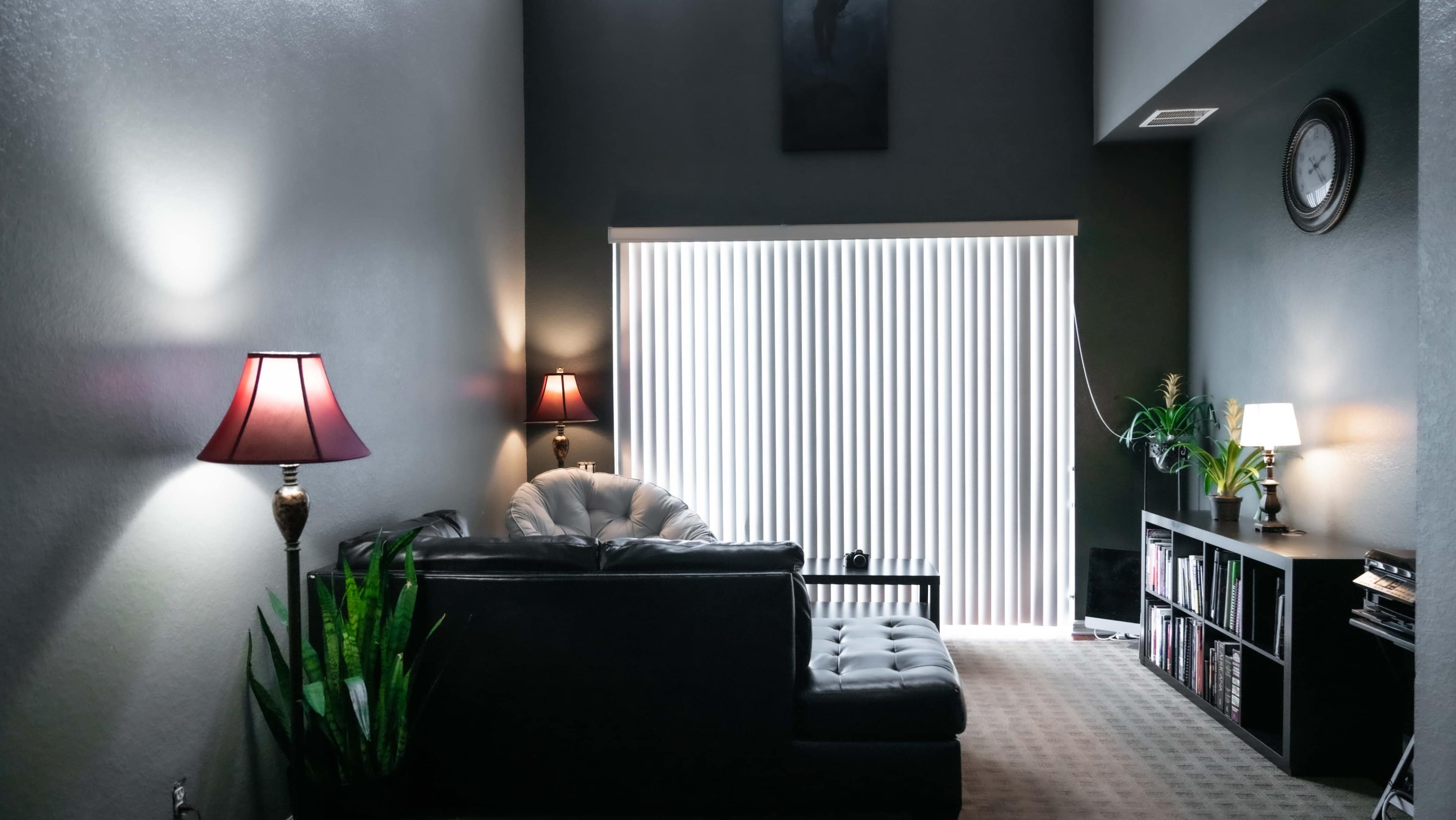 A modern living room with a sectional sofa, lamps, and a large window covered by vertical blinds.