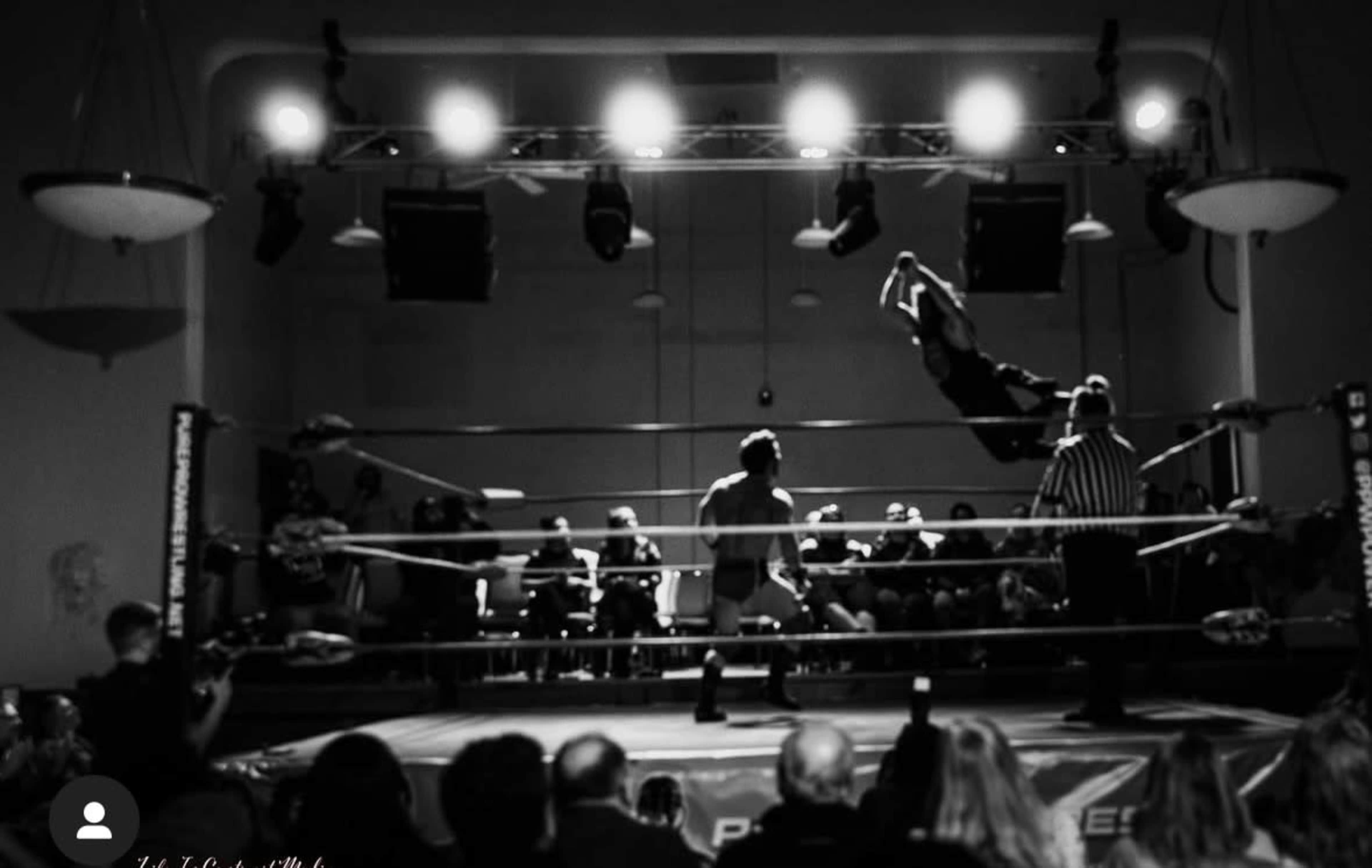 A wrestler leaps from the top rope toward another wrestler in a brightly lit ring while an audience watches from the sidelines.