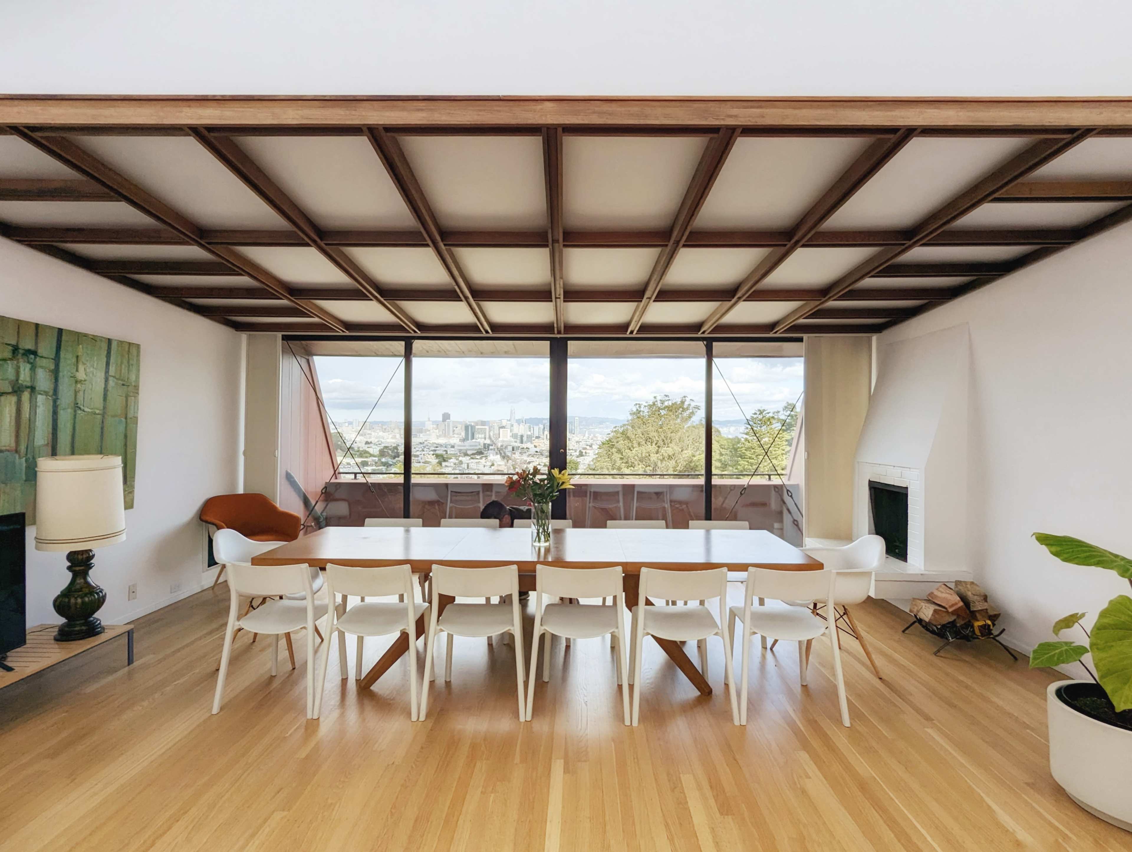 A spacious dining area features a long table surrounded by white chairs, with large glass windows offering a view of the city and mountains beyond.