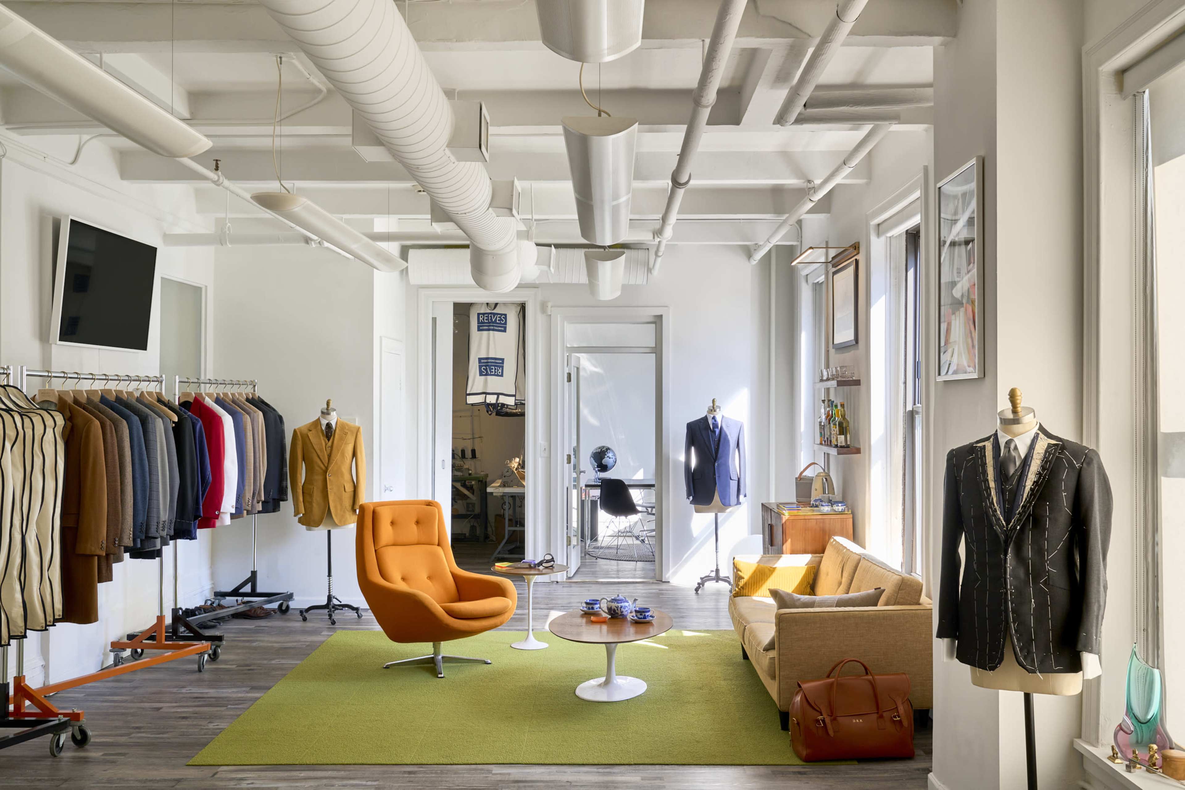 A modern studio space featuring a mix of clothing racks, an orange chair, a coffee table, and a sofa, with large windows allowing natural light to fill the room.