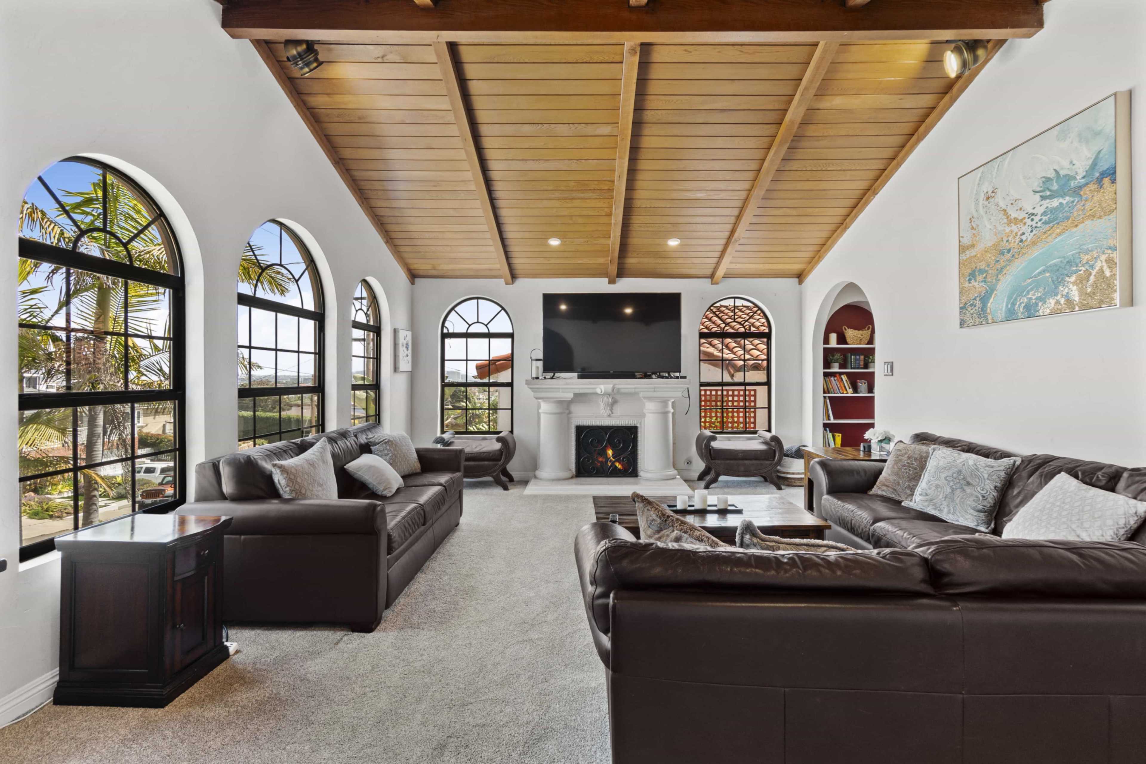 A spacious living room features dark leather sofas arranged around a coffee table, with large windows providing views of palm trees outside.