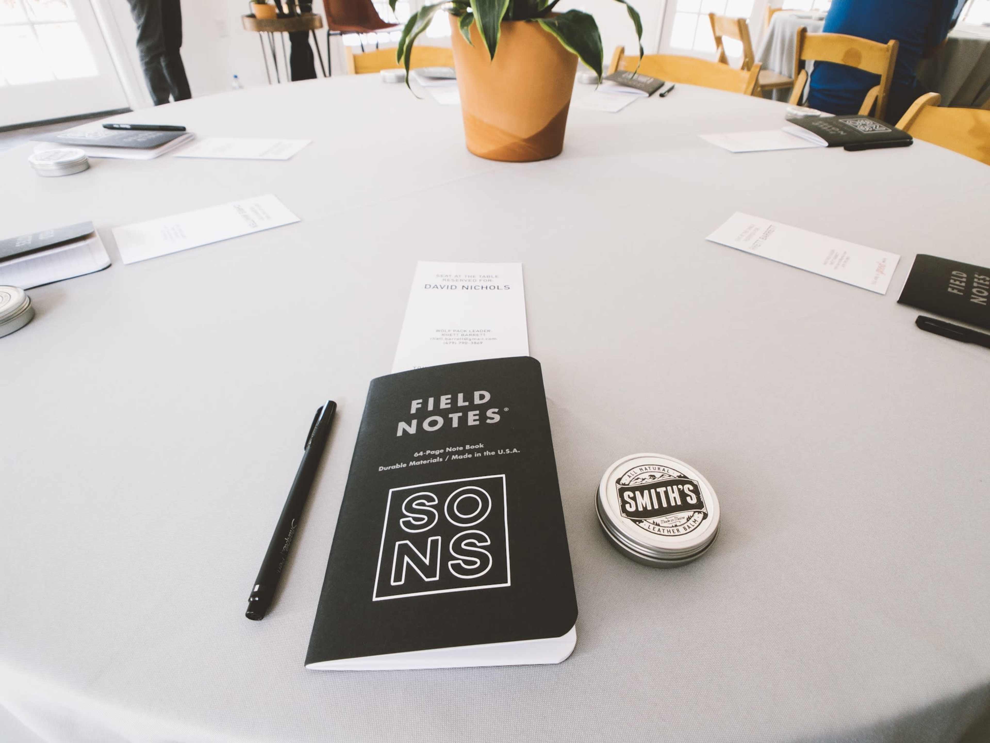 A table set for a meeting features a notebook labeled "FIELD NOTES," a pen, a circular tin labeled "SMITH'S," and printed materials arranged neatly.