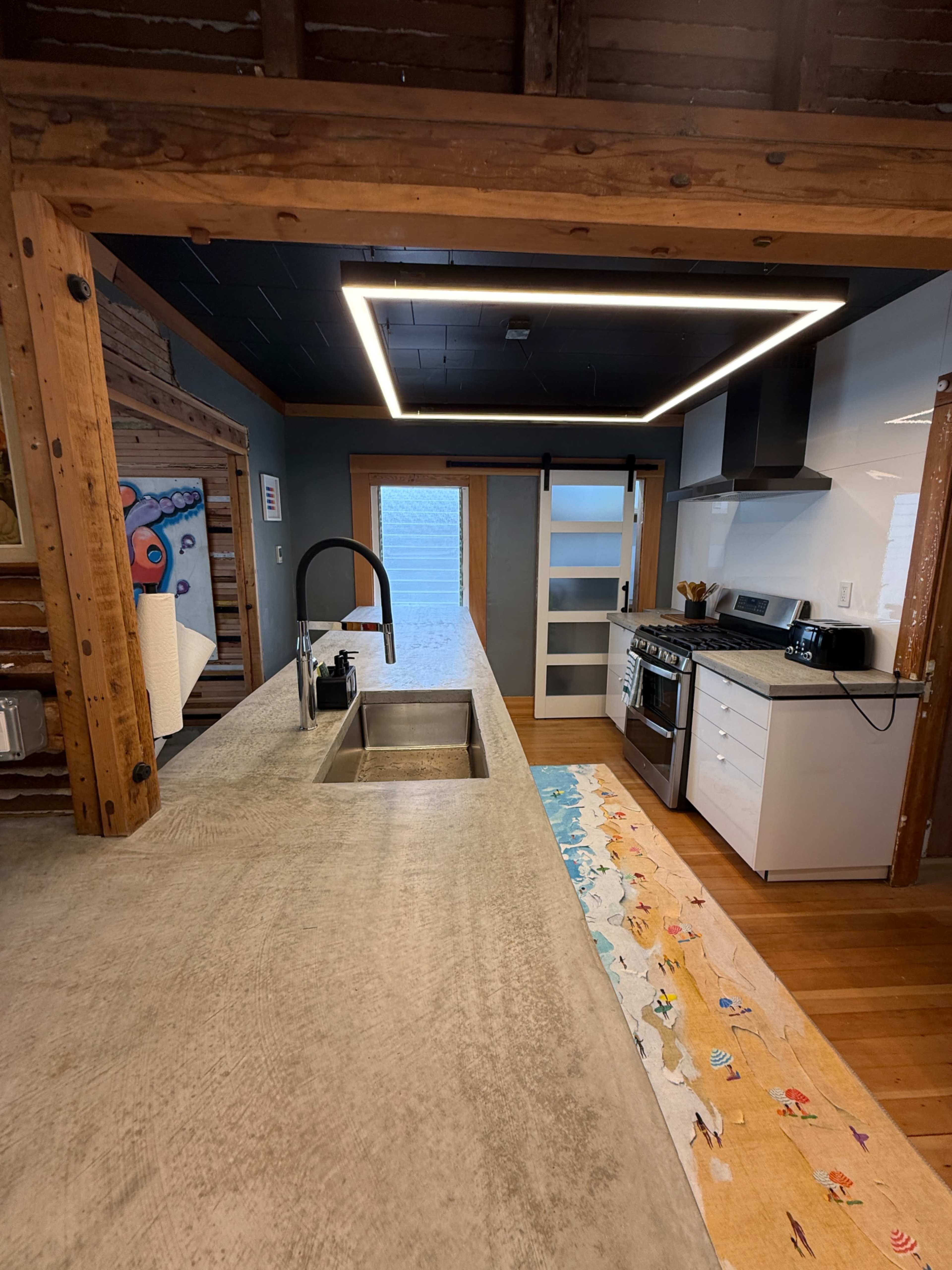 The image shows a modern kitchen with a concrete countertop, stainless steel appliances, and a wooden ceiling.