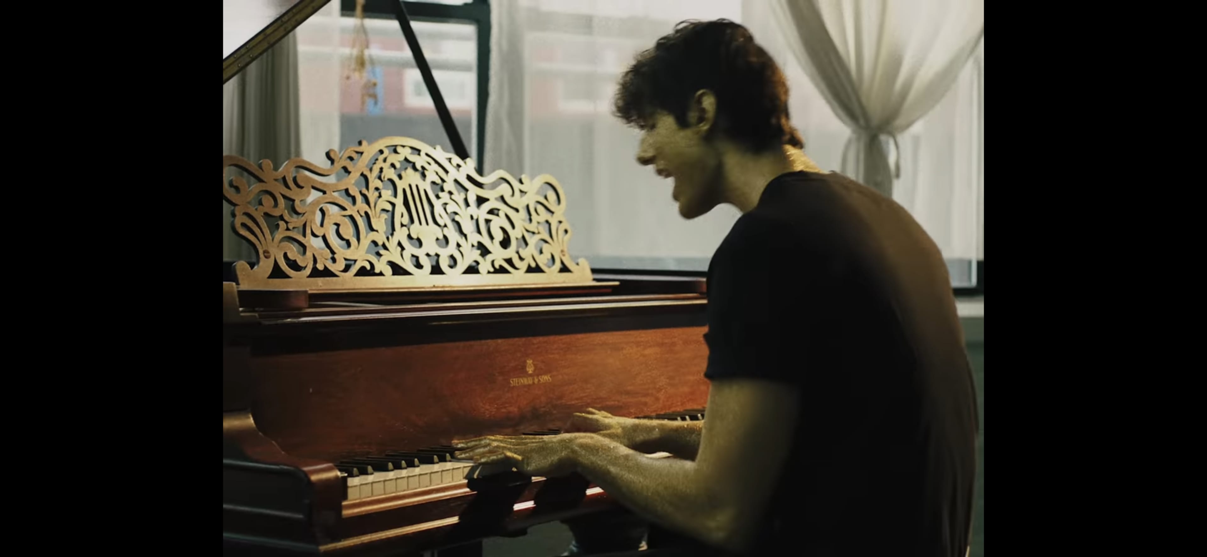 A young man is playing a wooden piano in a well-lit room with large windows.