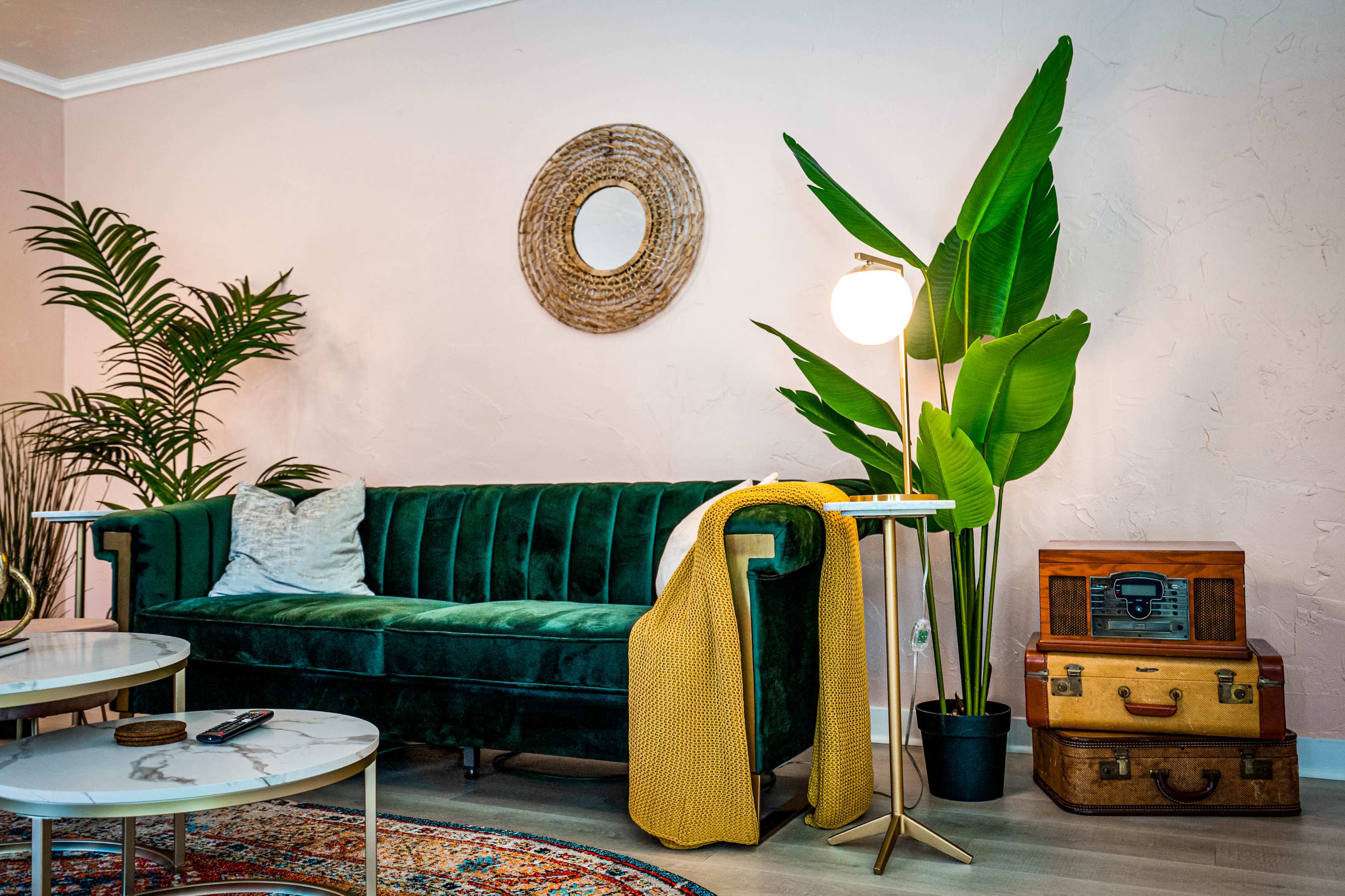 A green velvet sofa with a yellow throw blanket is positioned beside a tall plant, a round mirror, and a vintage trunk in a light-colored living room.