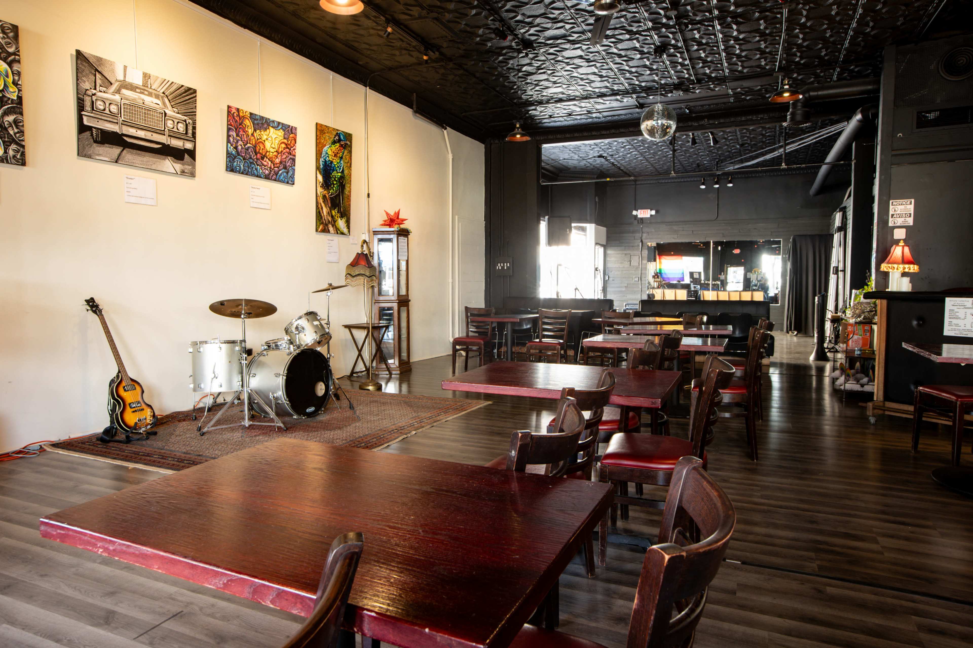The image shows an eclectic music venue featuring a drum set and guitar on display, surrounded by tables and artwork on the walls.