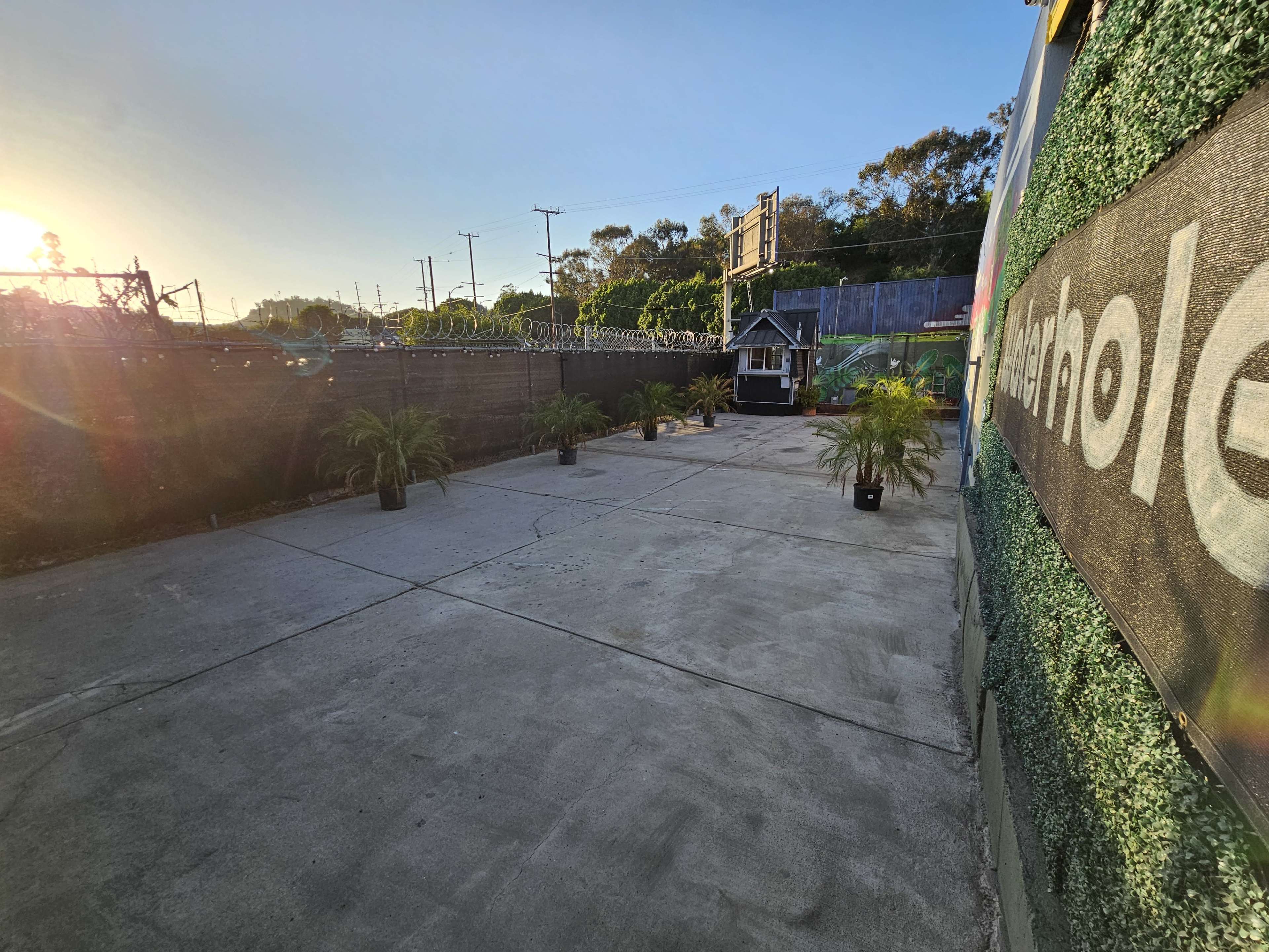An outdoor space with potted plants arranged on a concrete surface surrounded by a tall fence and a house structure in the background.